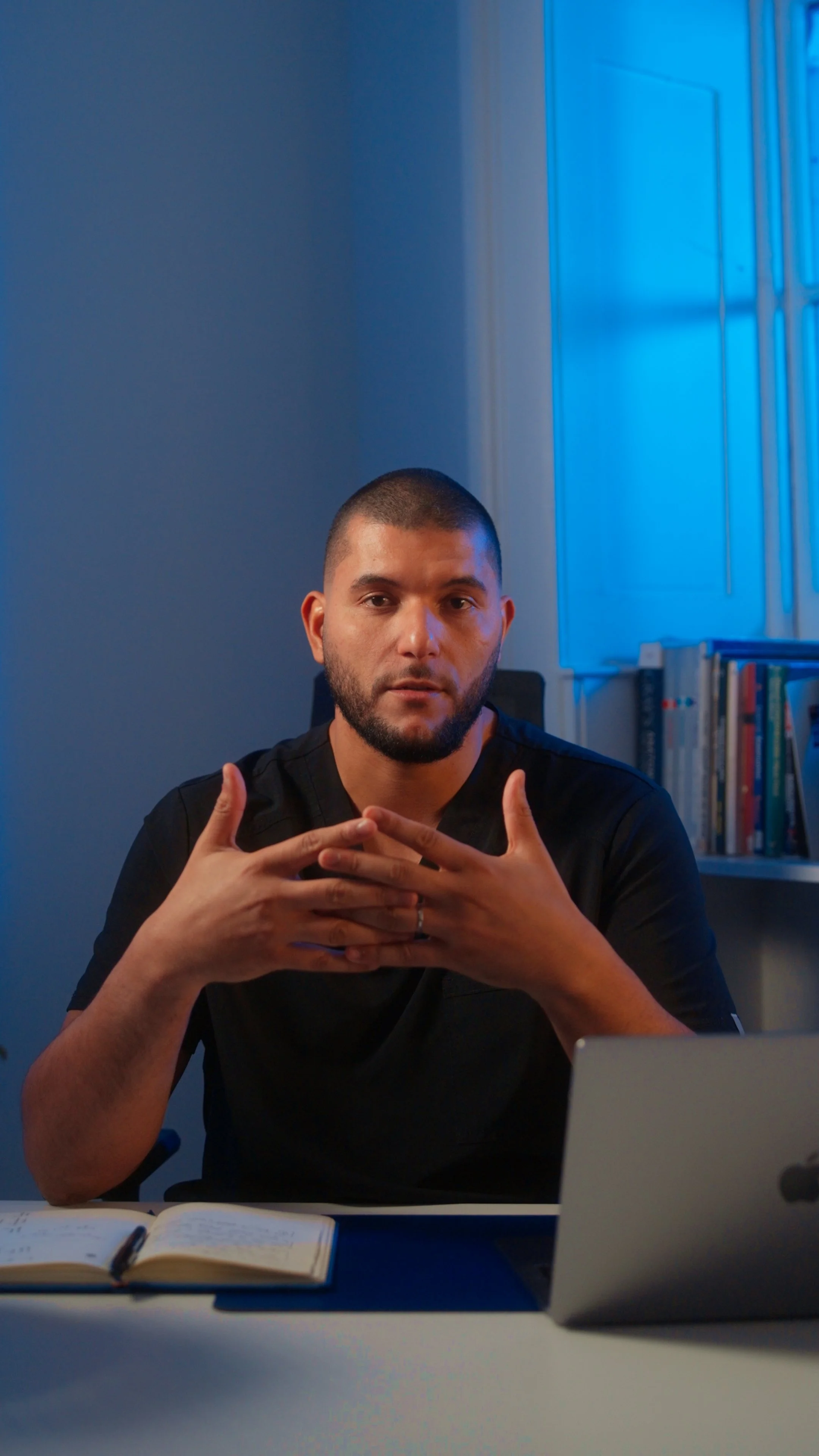 A man sitting at a desk with an open notebook and laptop, gesturing with his hands, in front of a bookshelf and a window with blue lighting.