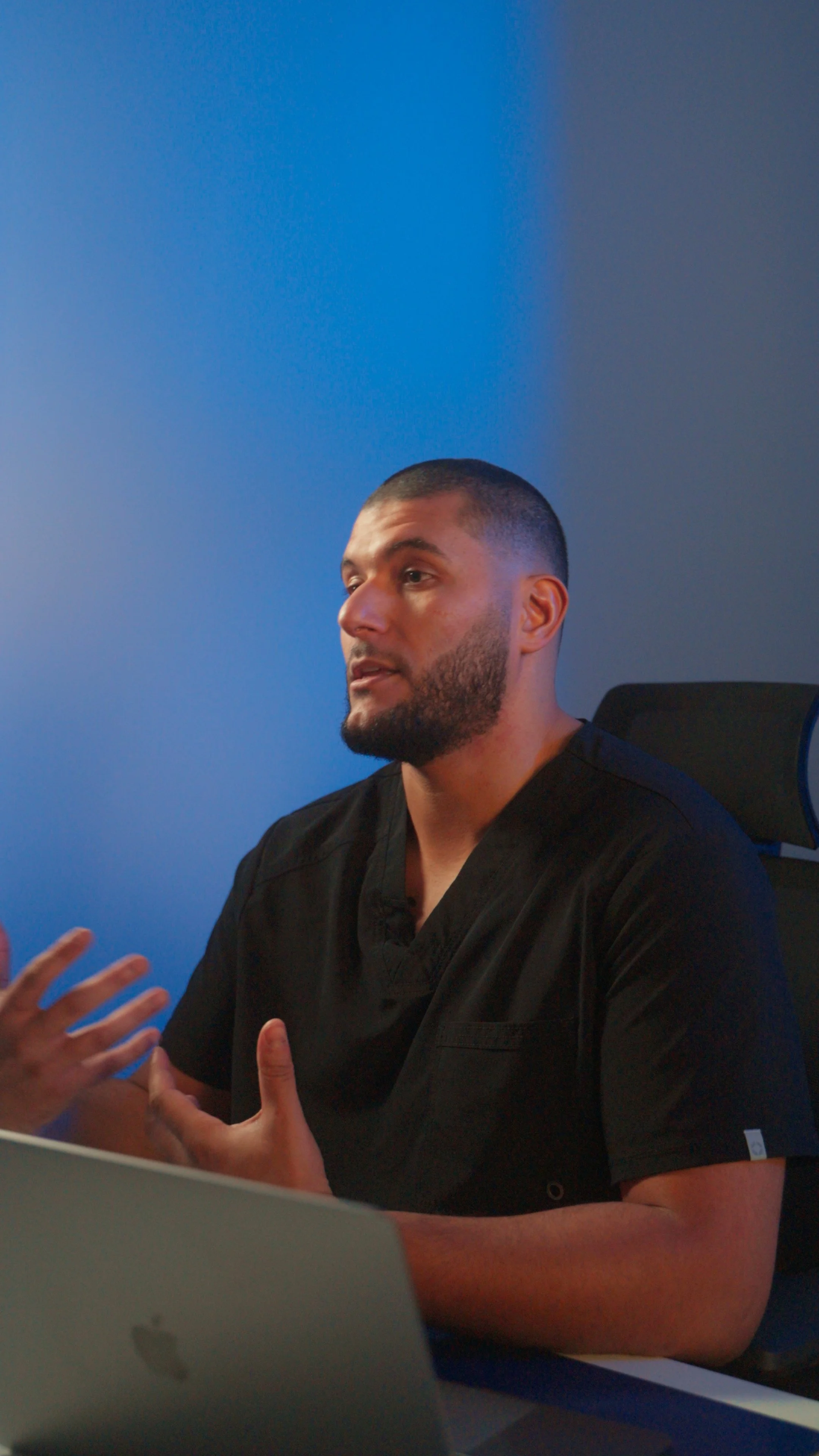 A man with short dark hair and a beard wearing a black top, engaged in conversation, gesturing with his hands. He is seated in front of a blue and gray background with part of a laptop in the foreground.