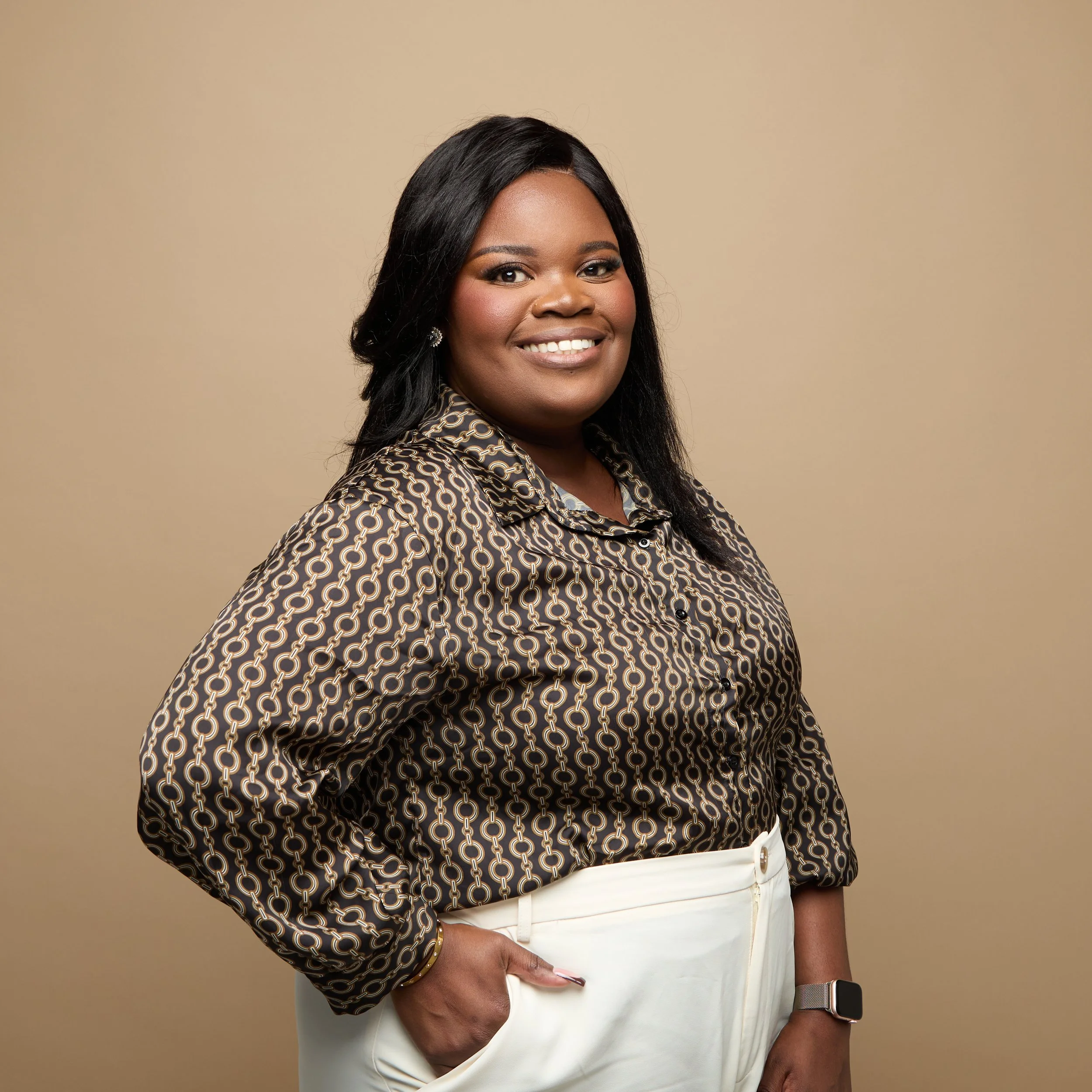 Une femme souriante avec des cheveux noirs longs, portant une chemise à motif de chaînes et un pantalon blanc, posant contre un fond beige.
