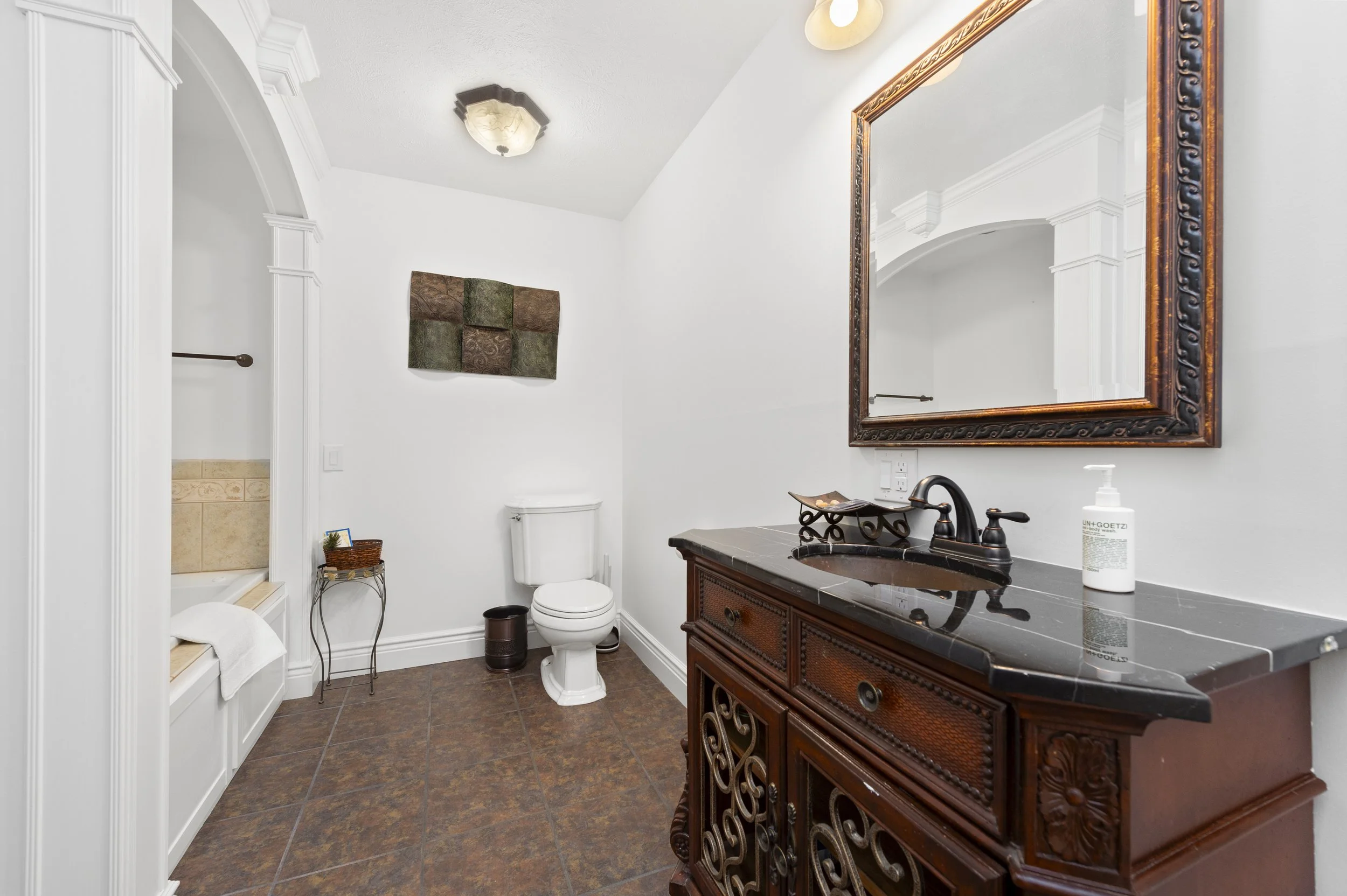 Elegant bathroom with a dark wooden vanity, black countertop, large decorative mirror, modern faucet, toilet, artwork on the wall, and tiled floor.