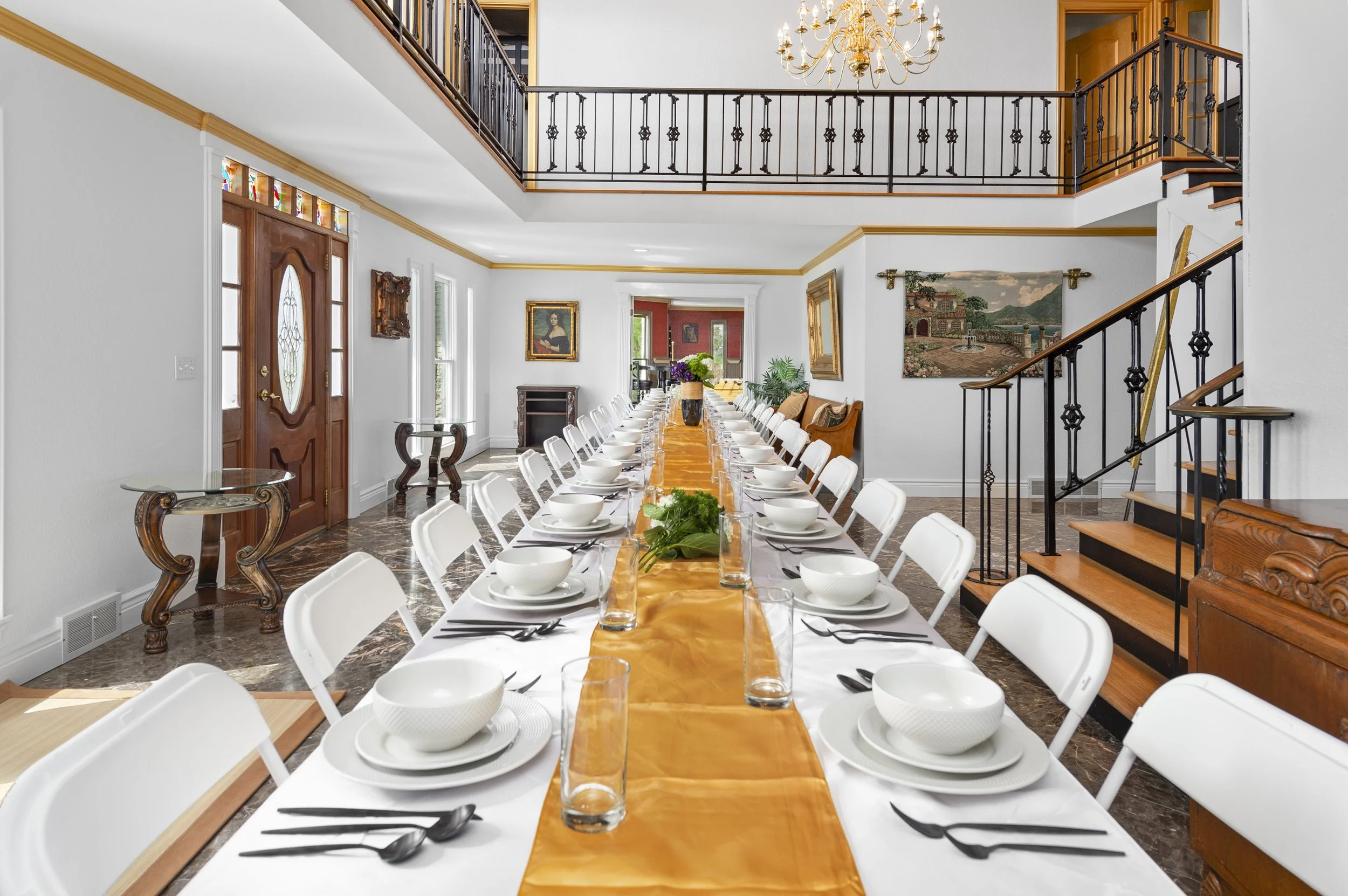 Elegant dining room with a long table set for a meal, featuring white chairs, gold table runner, plates, bowls, cutlery, and glasses. The room has marble flooring, decorative art, a staircase with iron railings, and a chandelier. A large entry door a