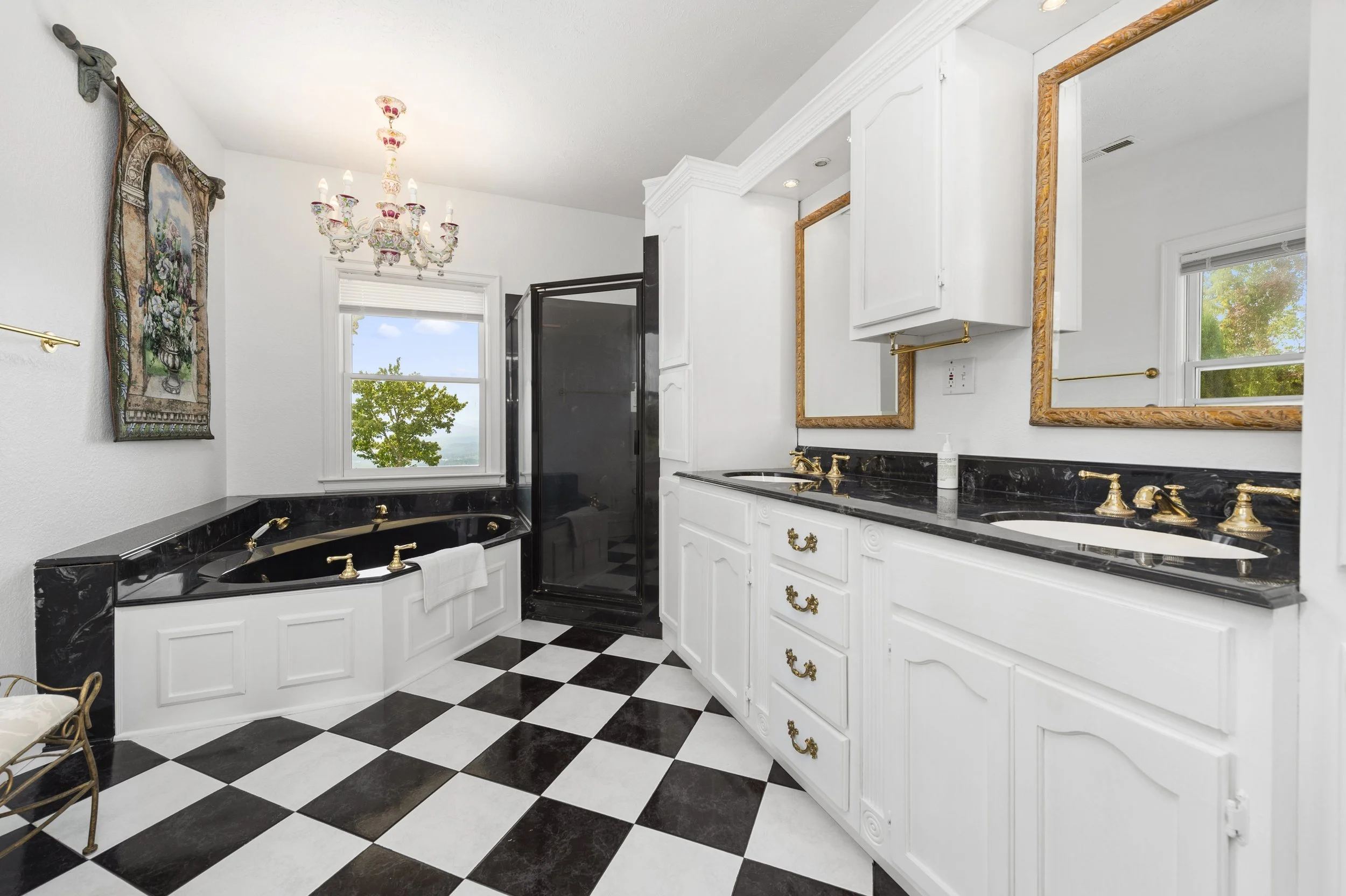 Elegant bathroom with black and white checkered floor, white cabinets with gold handles, black countertops, a black bathtub, gold faucets, a large wall mirror, and a decorative chandelier.