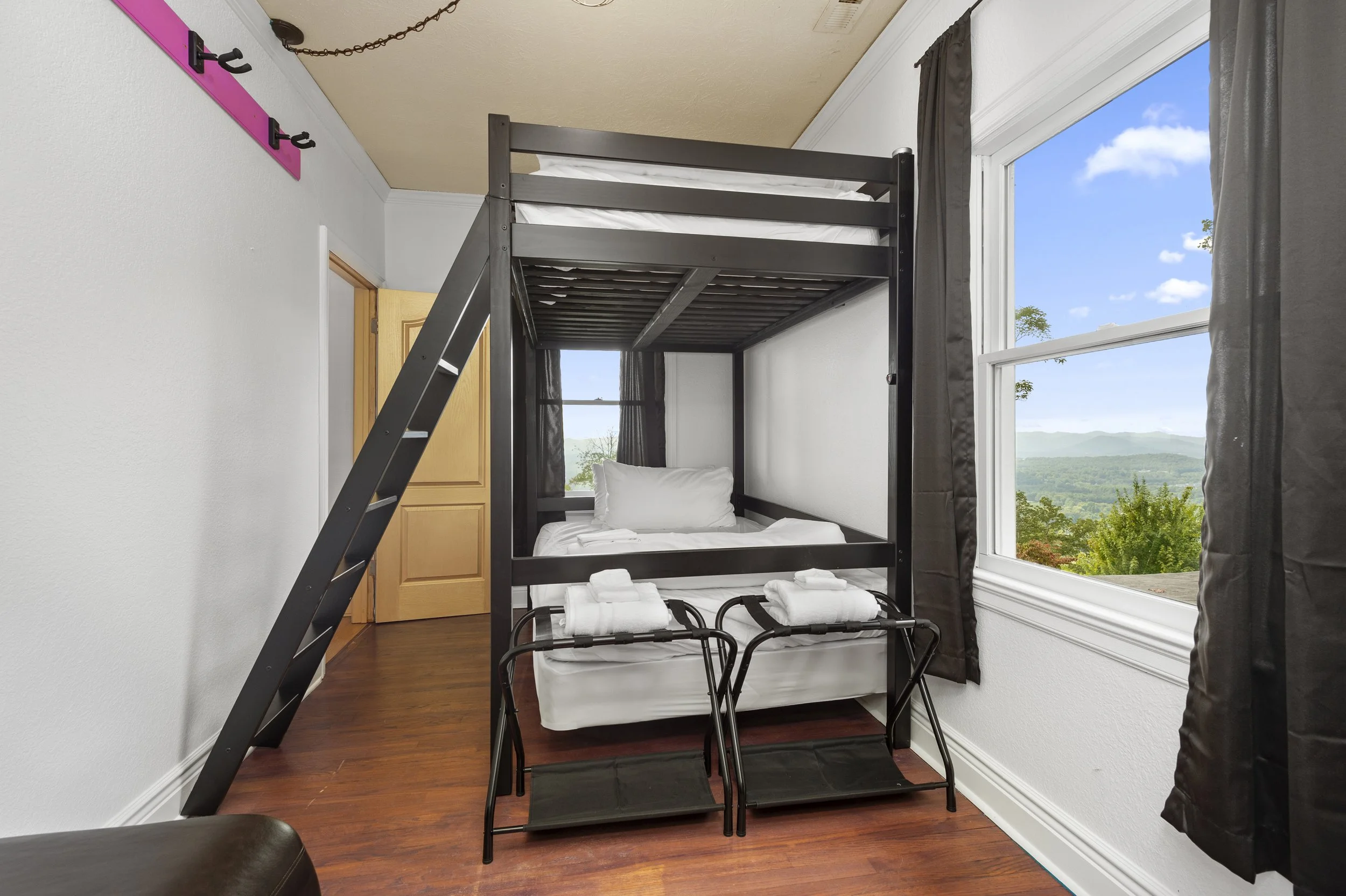 Room with bunk bed, ladder, and window view of mountains. Two luggage racks with towels are placed under the bed.