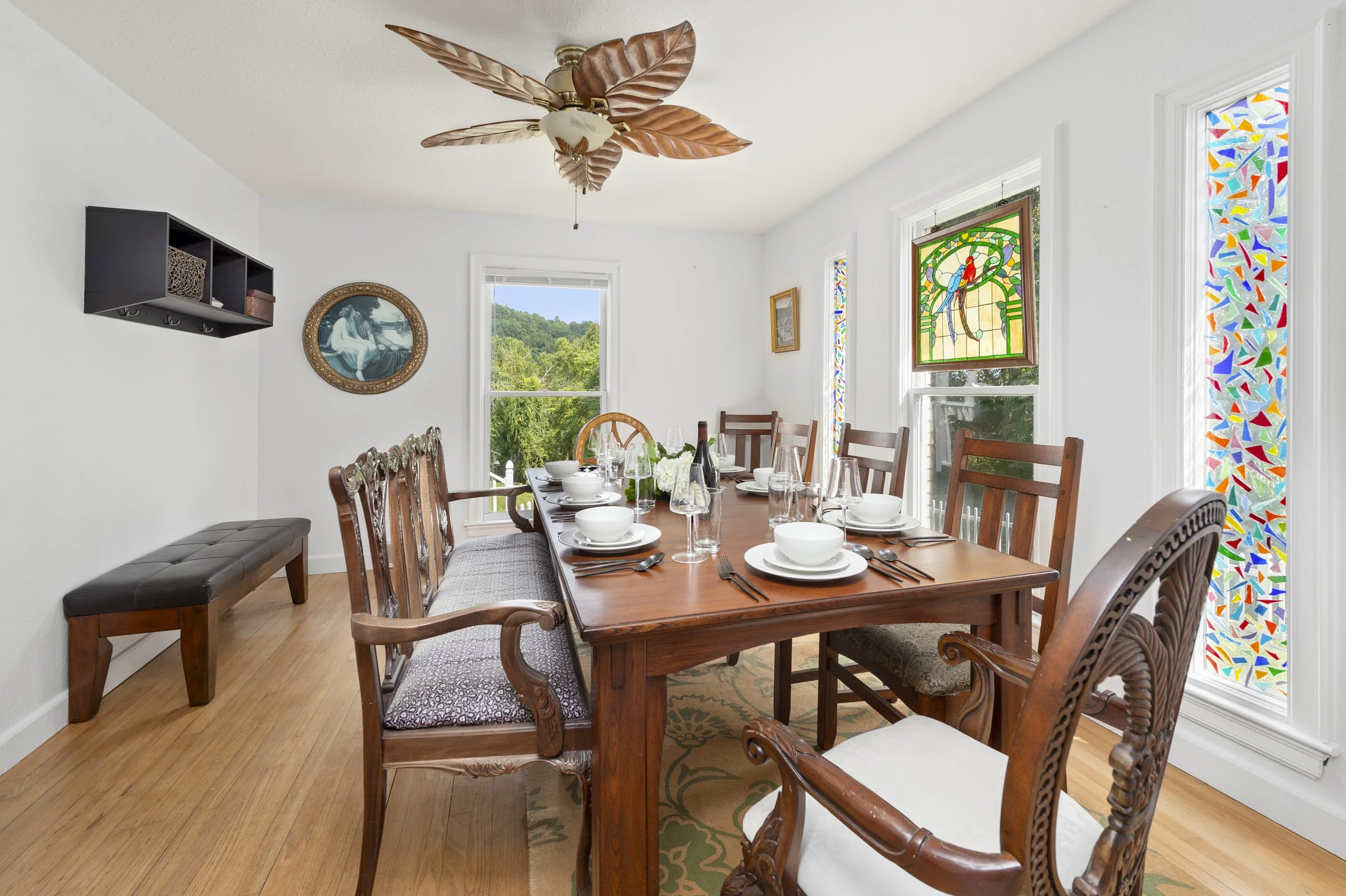 Dining room with wooden table and chairs, set for a meal, featuring a ceiling fan with leaf-shaped blades, stained glass windows, a wall mirror, and a patterned rug.