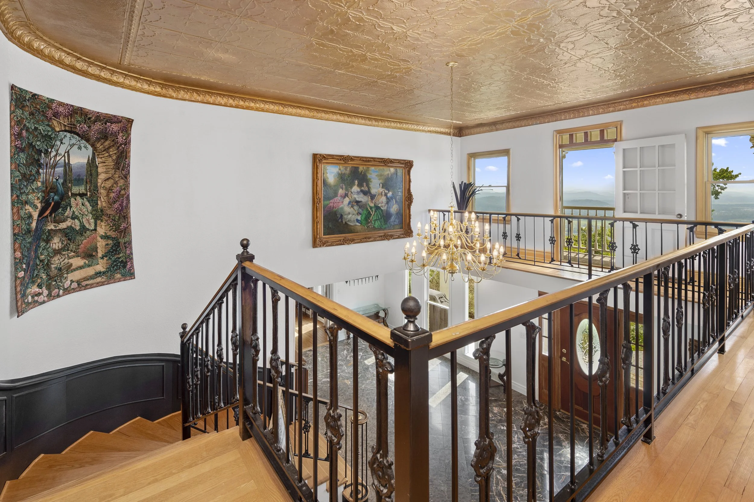 Elegant interior with wrought iron staircase, golden ceiling, chandelier, landscape painting, tapestry, and large windows with scenic view.