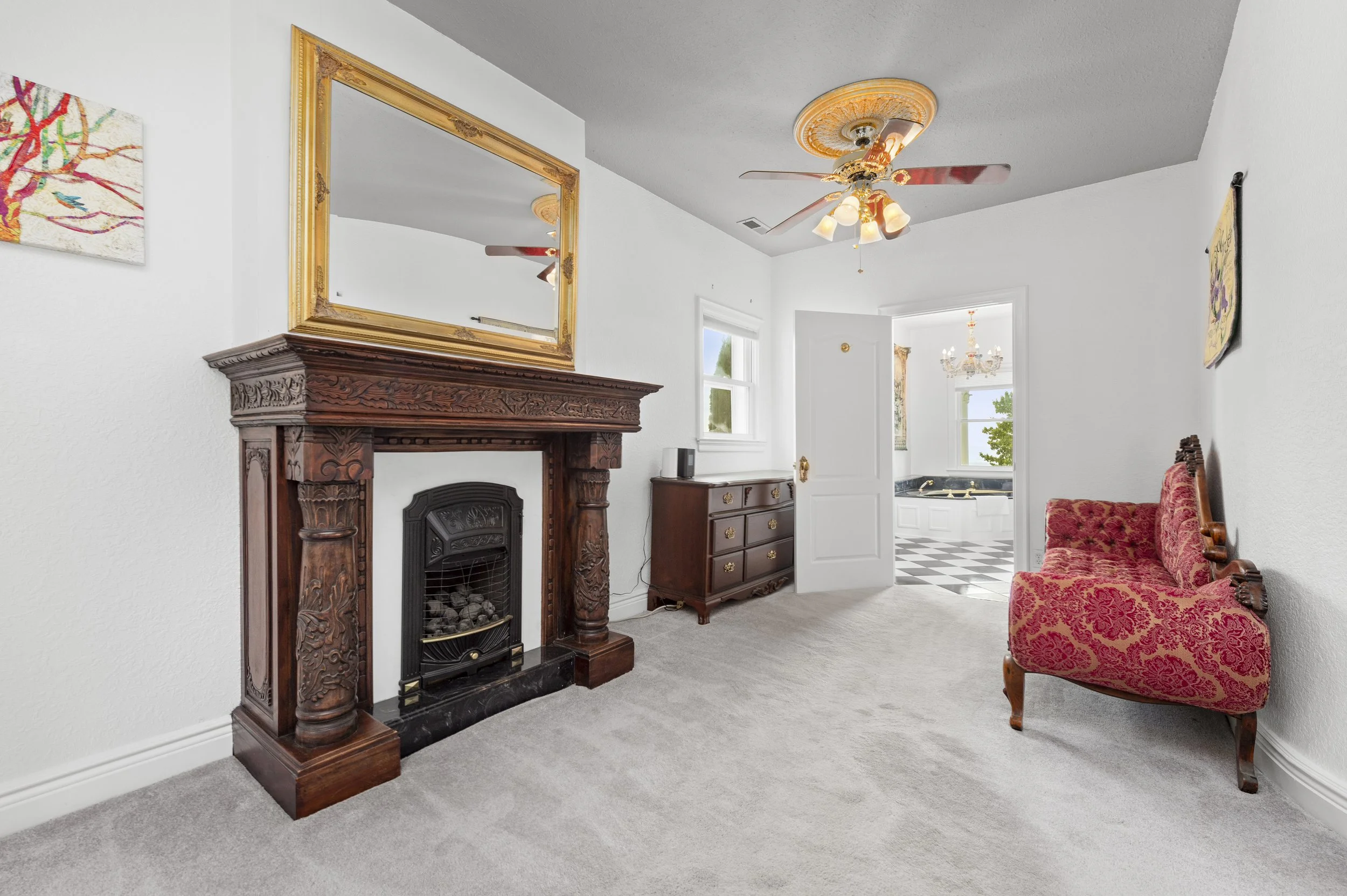 Elegant room with ornate wooden fireplace, large gold-framed mirror, red patterned settee, dark wood dresser, ceiling fan, and open door leading to bathroom with black and white checkered floor.