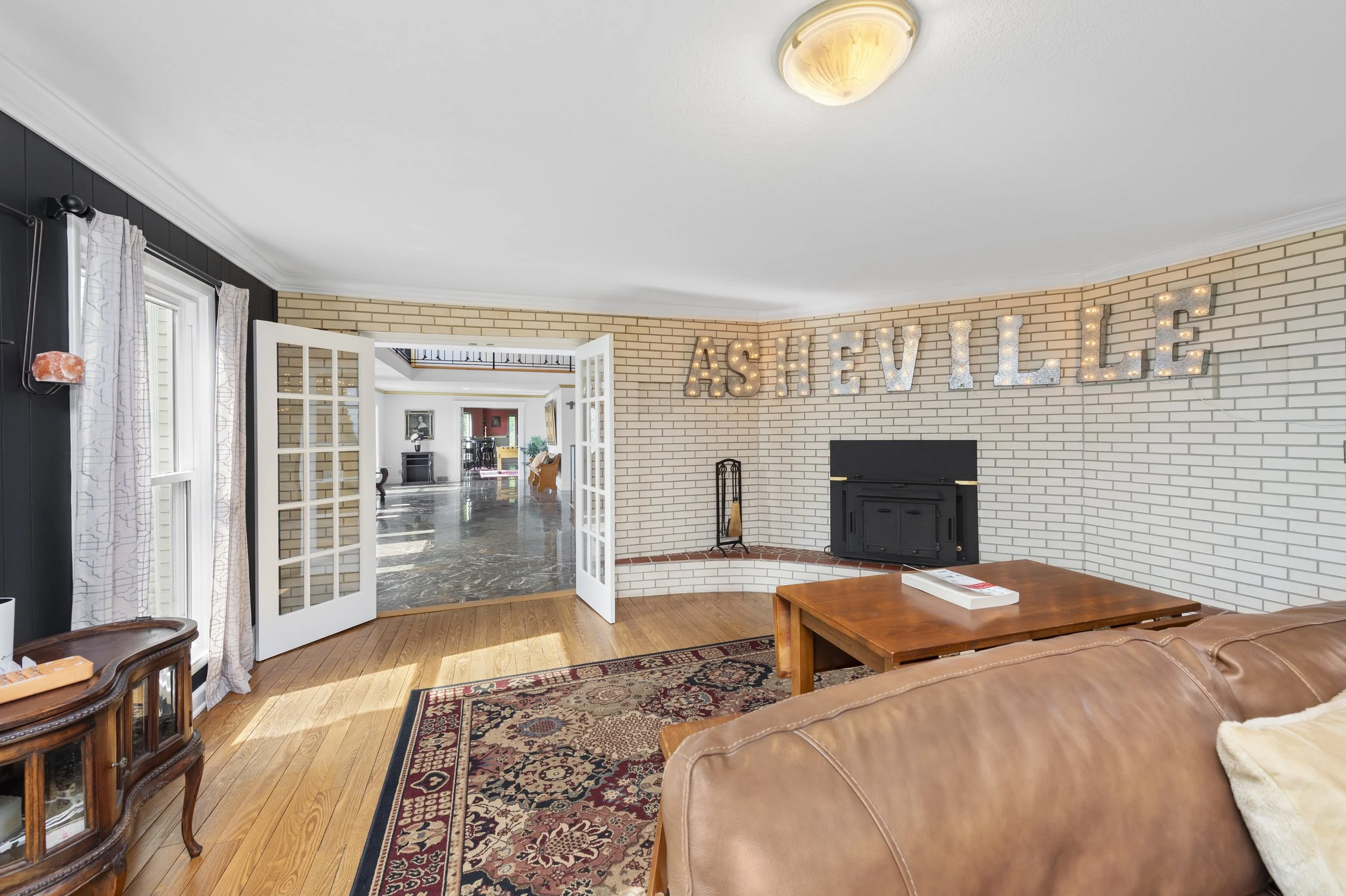 Living room with brick wall featuring "Asheville" in light-up letters, a fireplace, French doors, and wooden furniture.