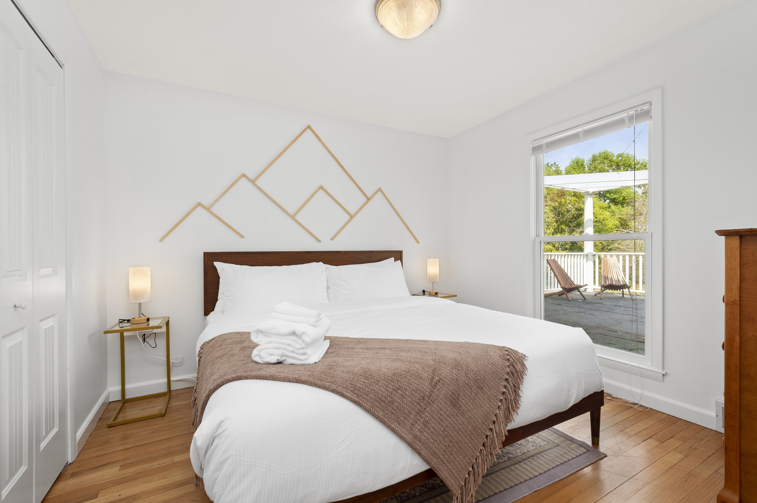 Modern bedroom with white bedding, a brown throw blanket, and wooden headboard. Geometric wall art above the bed. Two bedside tables with lamps. A large window shows a balcony with two chairs. Hardwood flooring and light fixture on the ceiling.