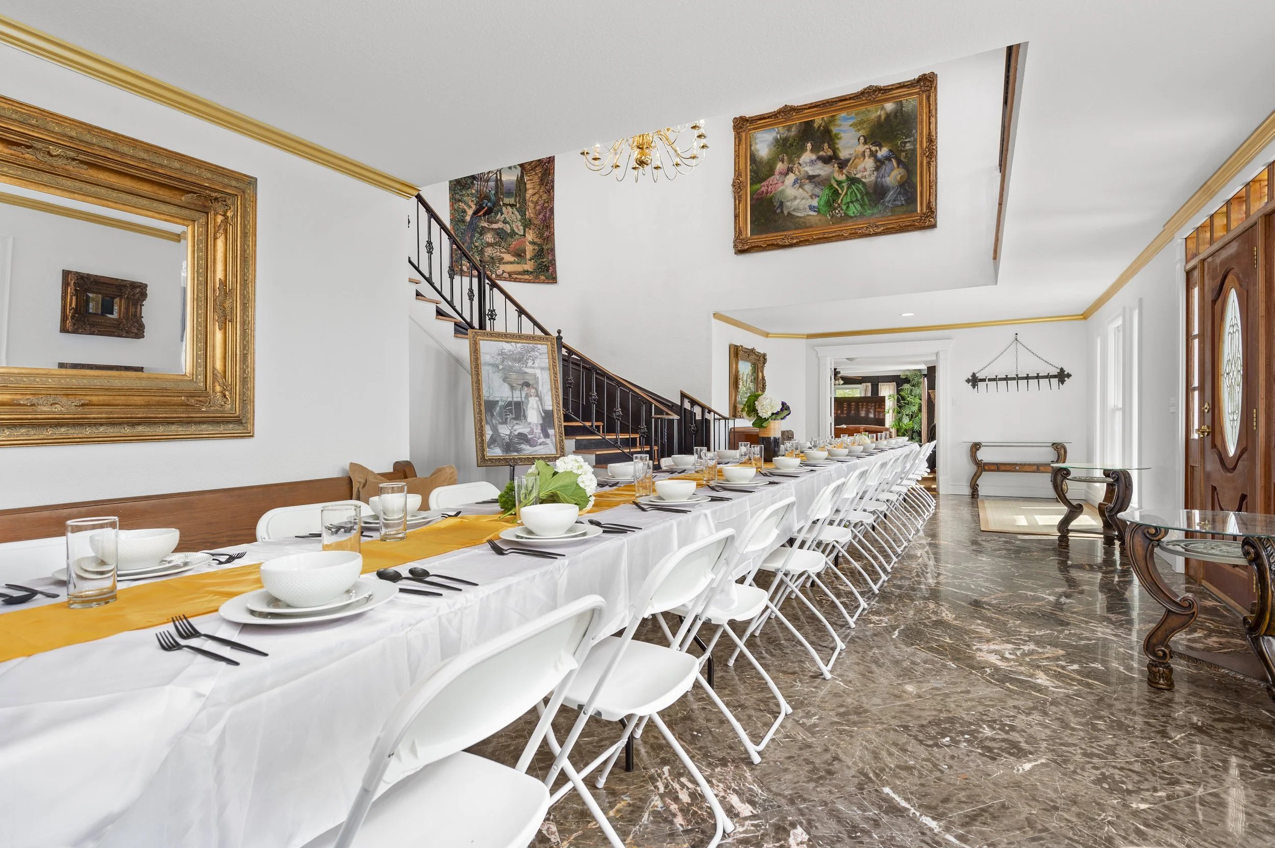 Elegant dining room with a long table set for a meal, featuring white folding chairs, tableware, and a yellow table runner. The room has a large mirror, framed paintings, and a staircase leading upstairs.