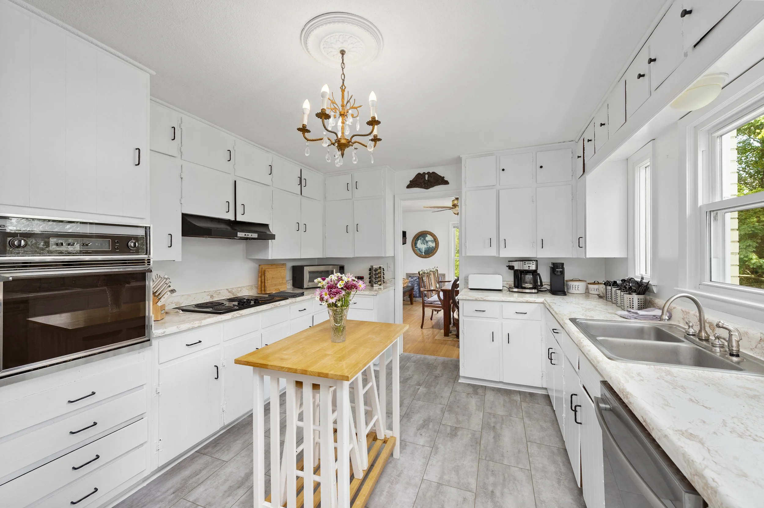 A spacious white kitchen with marble countertops, a stainless steel sink, gas stovetop, and a built-in oven. There's a small wooden island with a vase of flowers. Overhead, a chandelier hangs. Large windows provide natural light. To the side, there's
