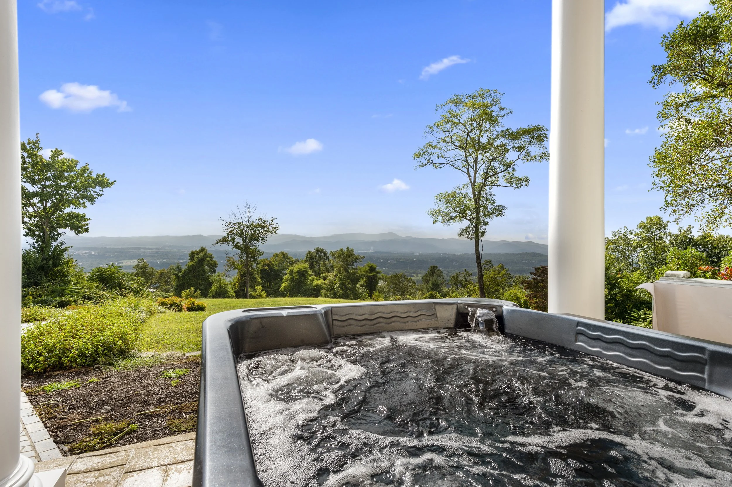 Hot tub on a patio overlooking a scenic landscape with trees and distant mountains under a clear blue sky.