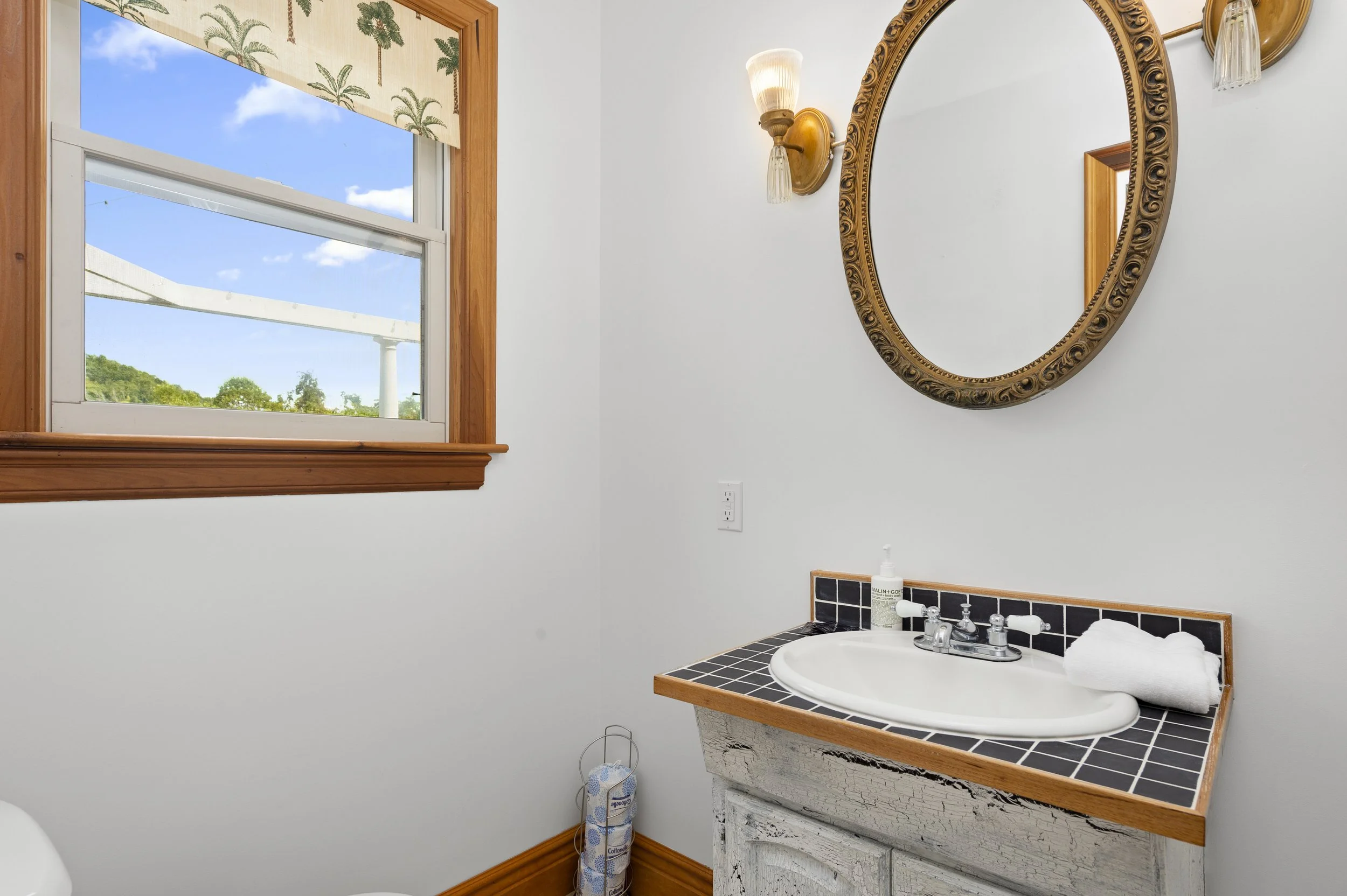 Bathroom with a window view, oval mirror, wall sconce, tiled countertop sink, and toilet paper holder.