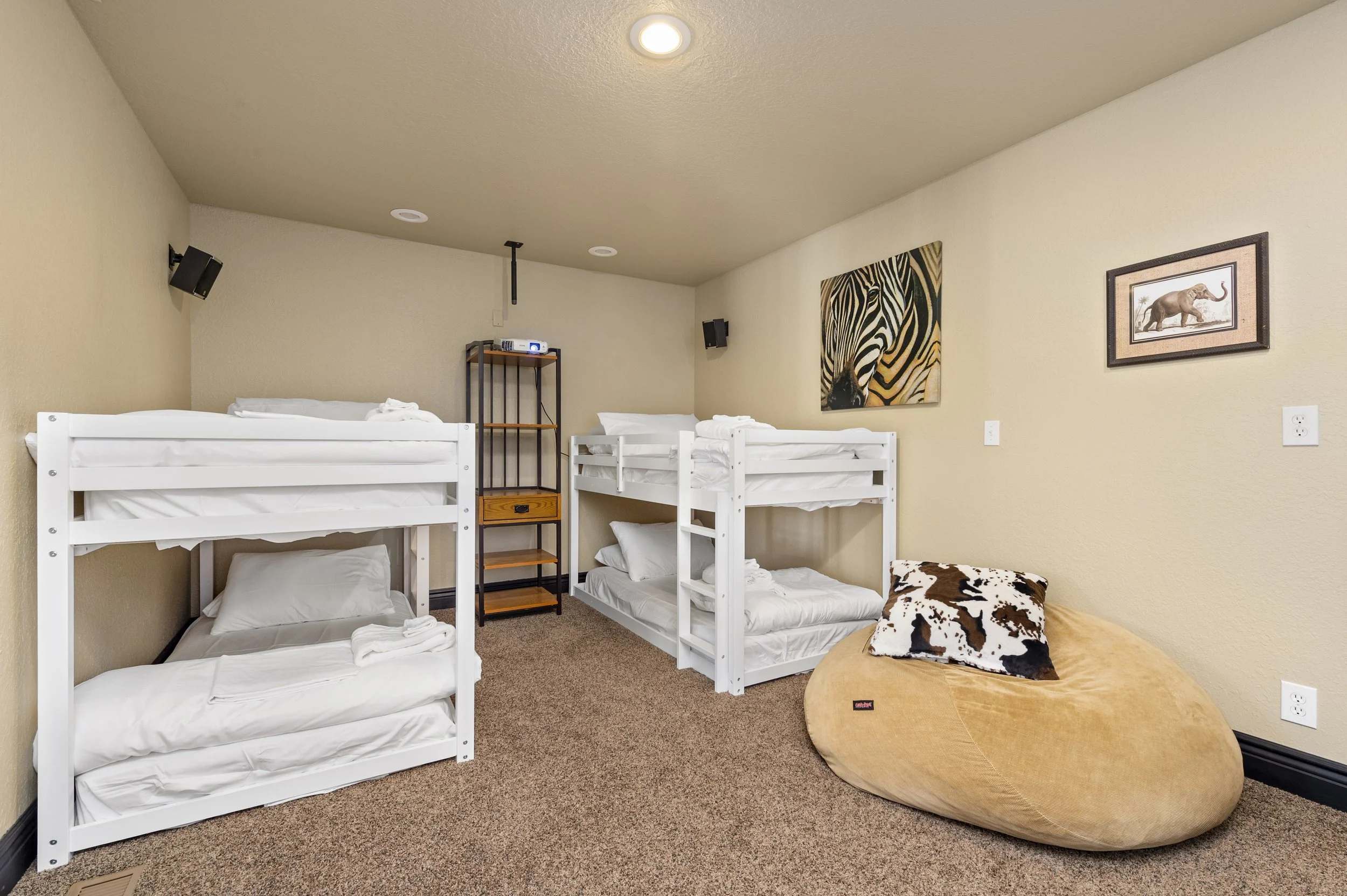 Room with two sets of white bunk beds, a bean bag with a cow print pillow, a shelving unit, and zebra artwork on beige walls.