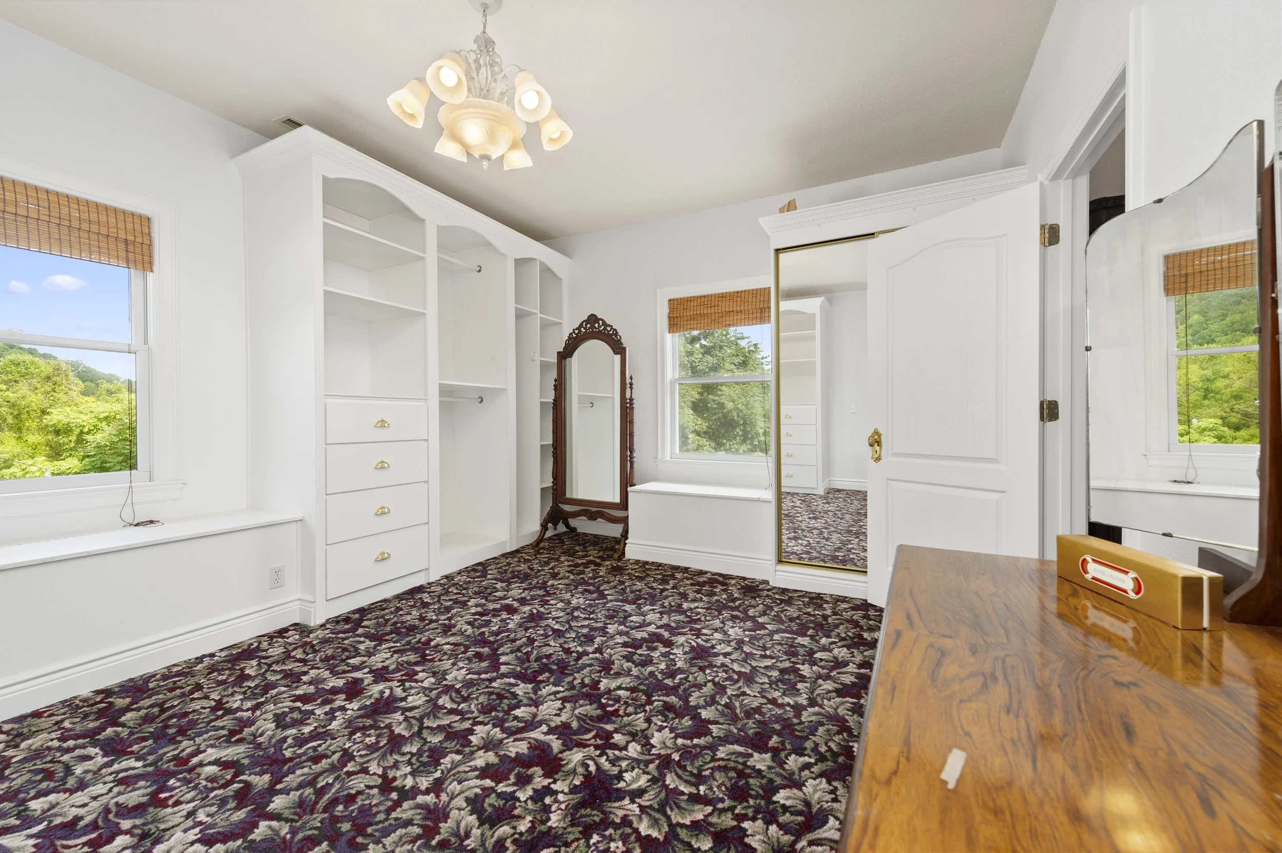 Interior of a white closet room with built-in shelves, floral carpet, standing mirror, and a chandelier.