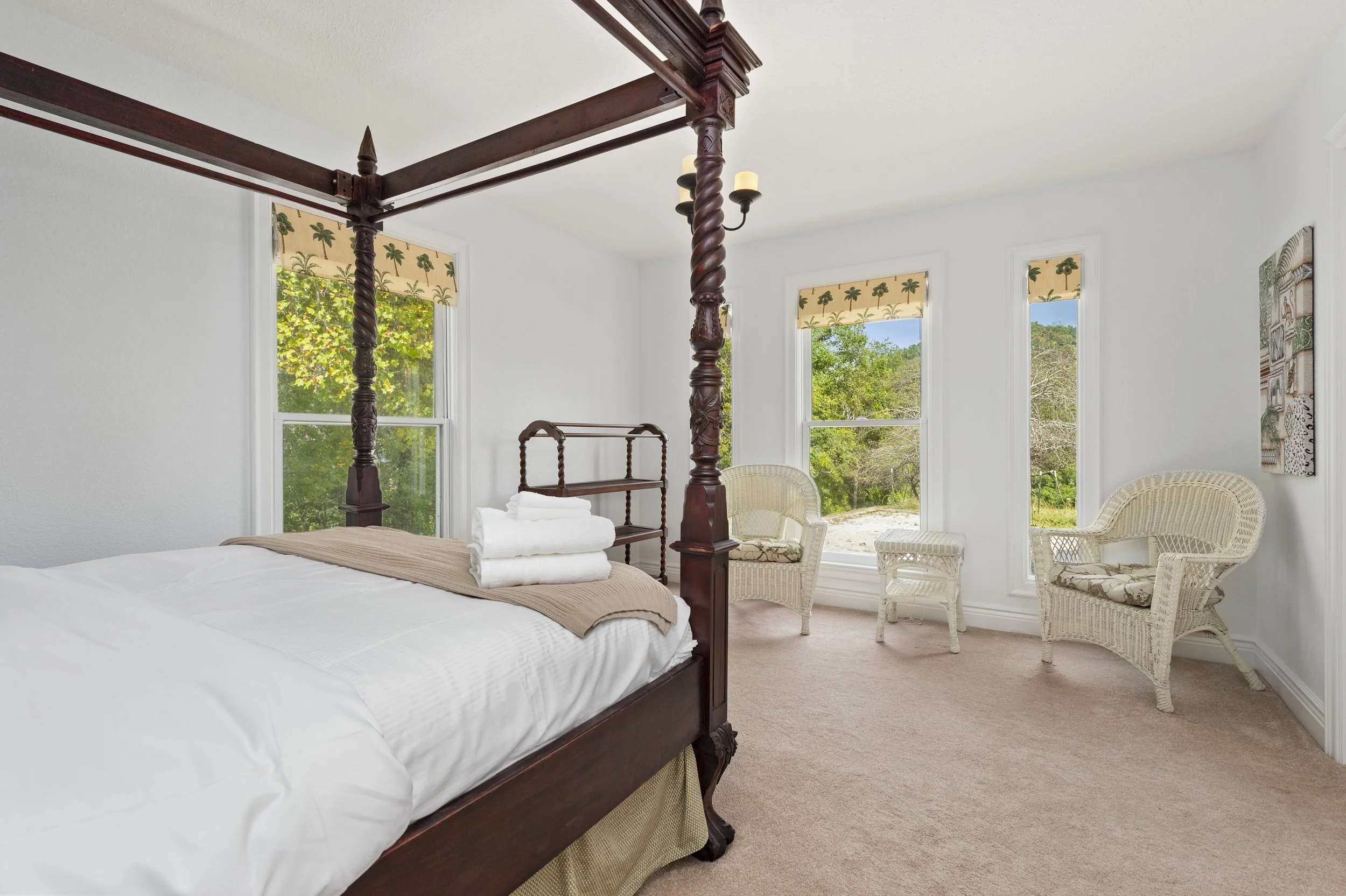 Bedroom with a four-poster bed and wicker furniture, including two chairs and a small table by large windows with palm tree curtains.