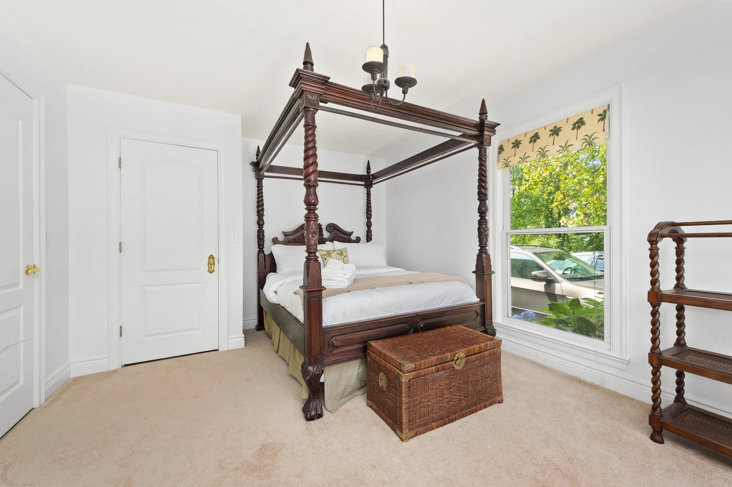 Bedroom with a four-poster bed, wicker chest, wooden shelves, and window with view of greenery and a car.