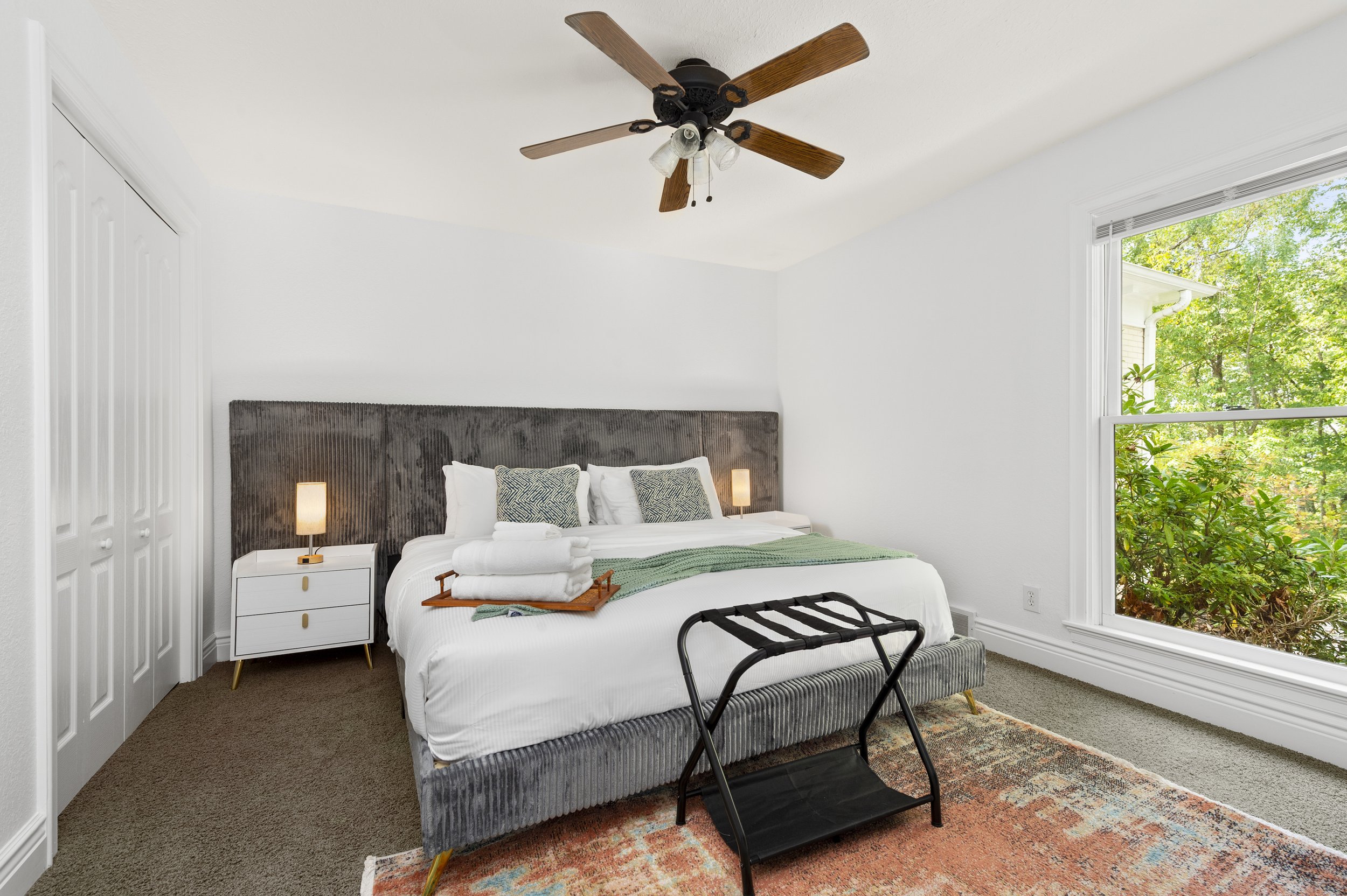 Modern bedroom with large bed, gray headboard, and decorative pillows. Bedside table with lamp, ceiling fan, and large window with view of green foliage. Tray with towels on bed, luggage rack on floor, and colorful rug.