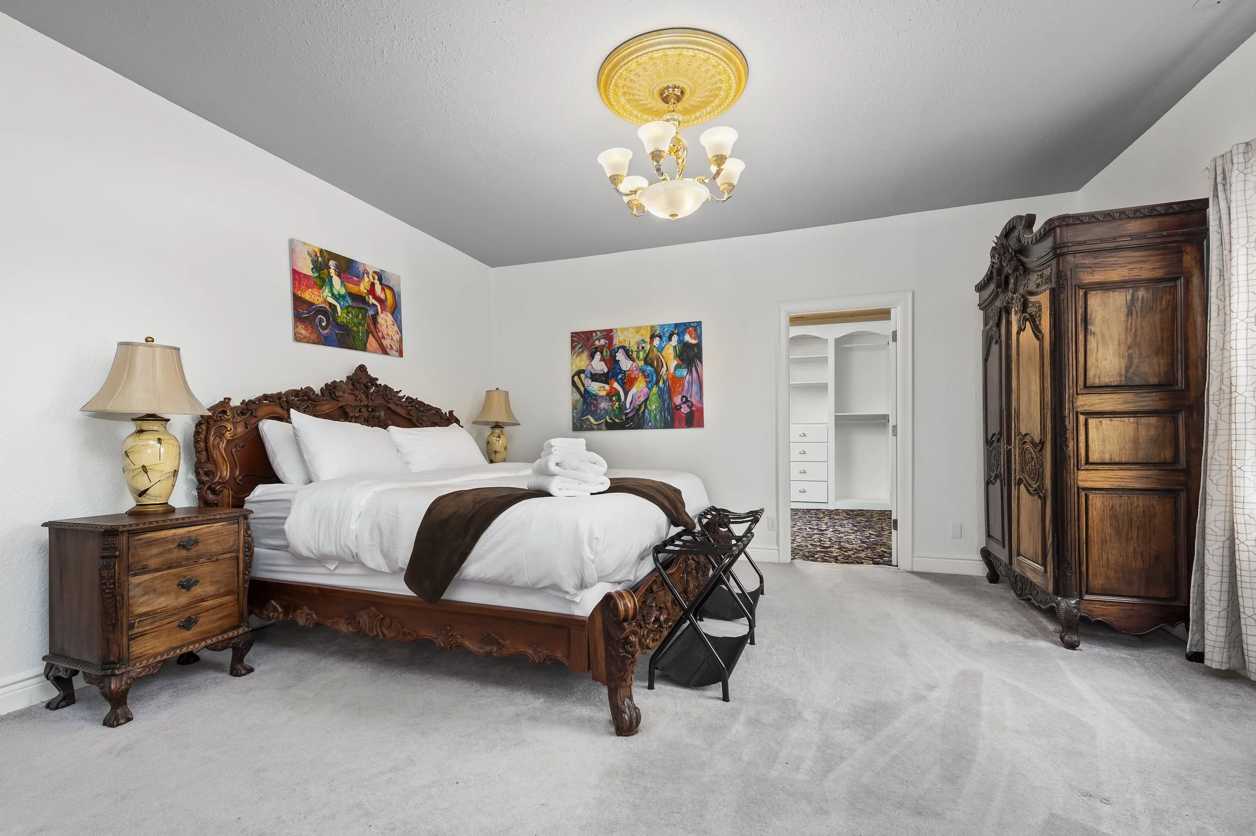 Elegant bedroom with ornate wooden furniture, including a bed, nightstand, and armoire. The bed is made with white linens and a dark brown throw. Two colorful paintings adorn the walls. A ceiling light fixture is visible, and there's a view into a cl