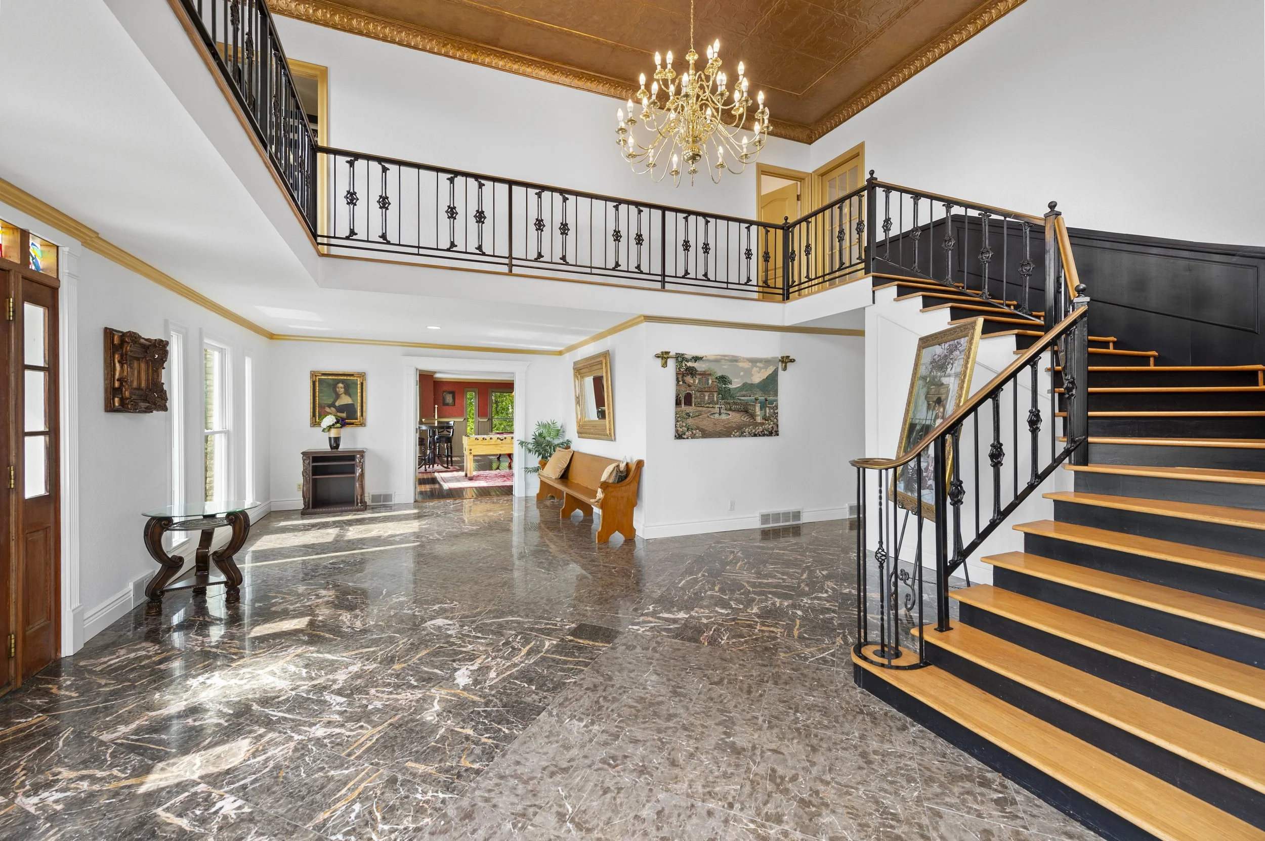 Elegant entryway with marble flooring, ornate chandelier, wooden staircase, and decorative railing. Walls adorned with framed artwork, a console table, and wooden bench. Bright natural light floods through large windows.