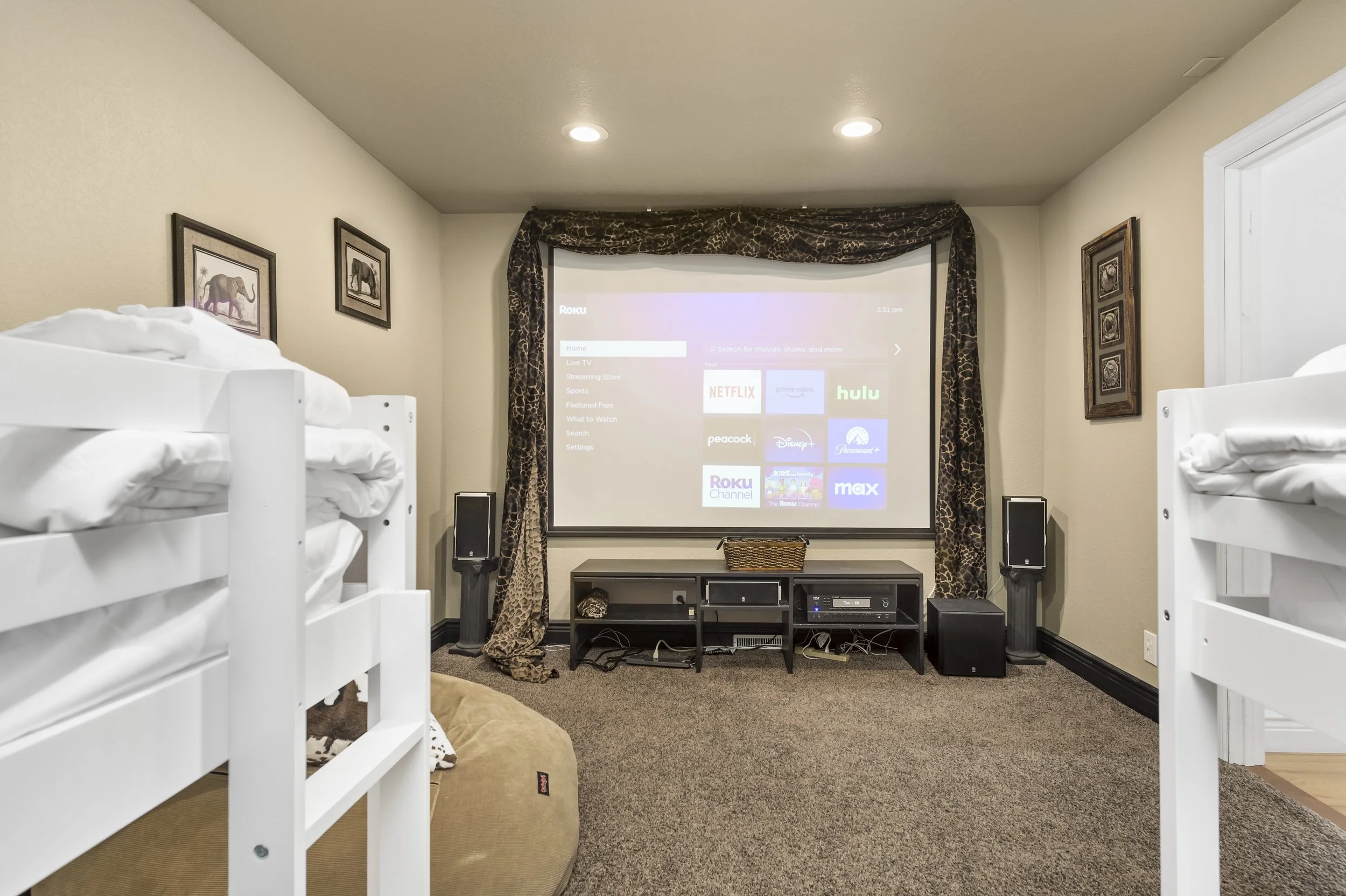 A home theater room with a large projection screen displaying streaming apps, flanked by speakers. White bunk beds are on each side, and framed art decorates the walls.