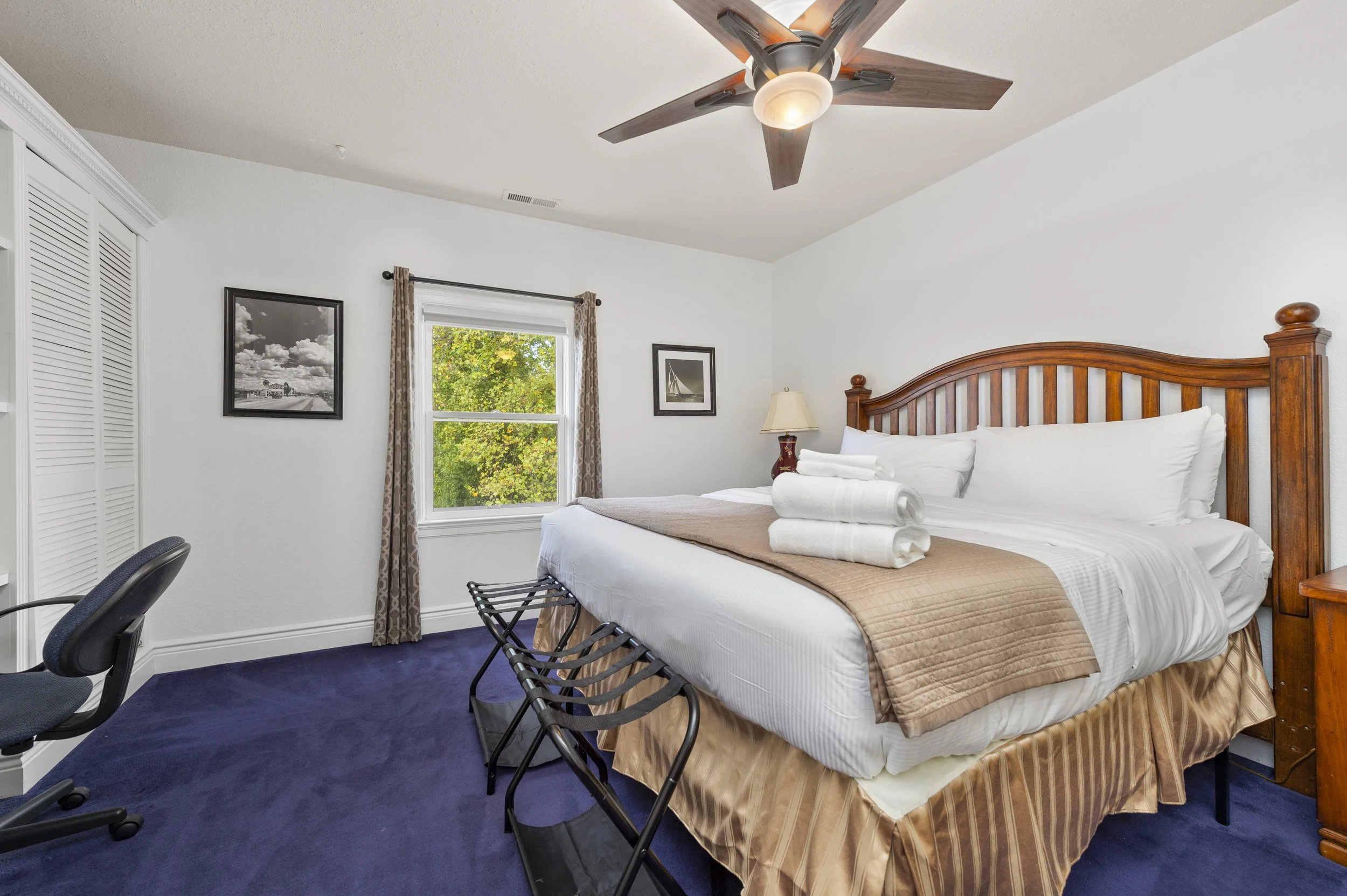 A bedroom with a wooden headboard bed, white linens, and folded towels on top. A ceiling fan is overhead, and there's a window with curtains showing green trees outside. A desk chair sits in the corner, and the floor is carpeted in dark blue.