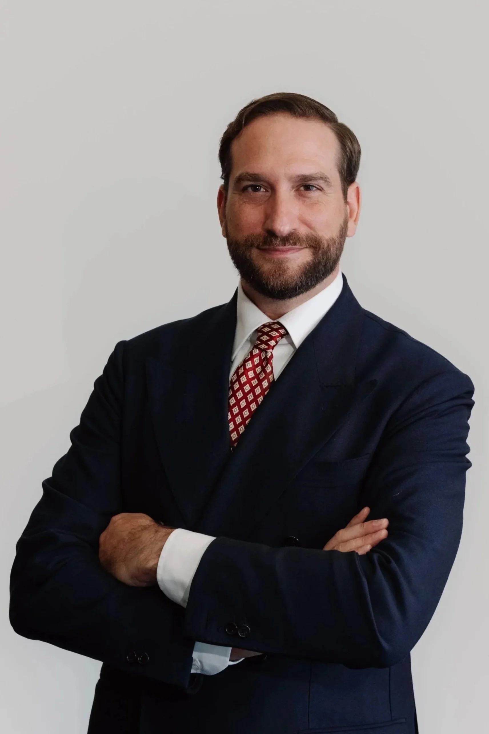 Uomo con barbe, in giacca blu, camicia bianca e cravatta di motivo geometrico, con braccia incrociate, sorridente, sfondo bianco.