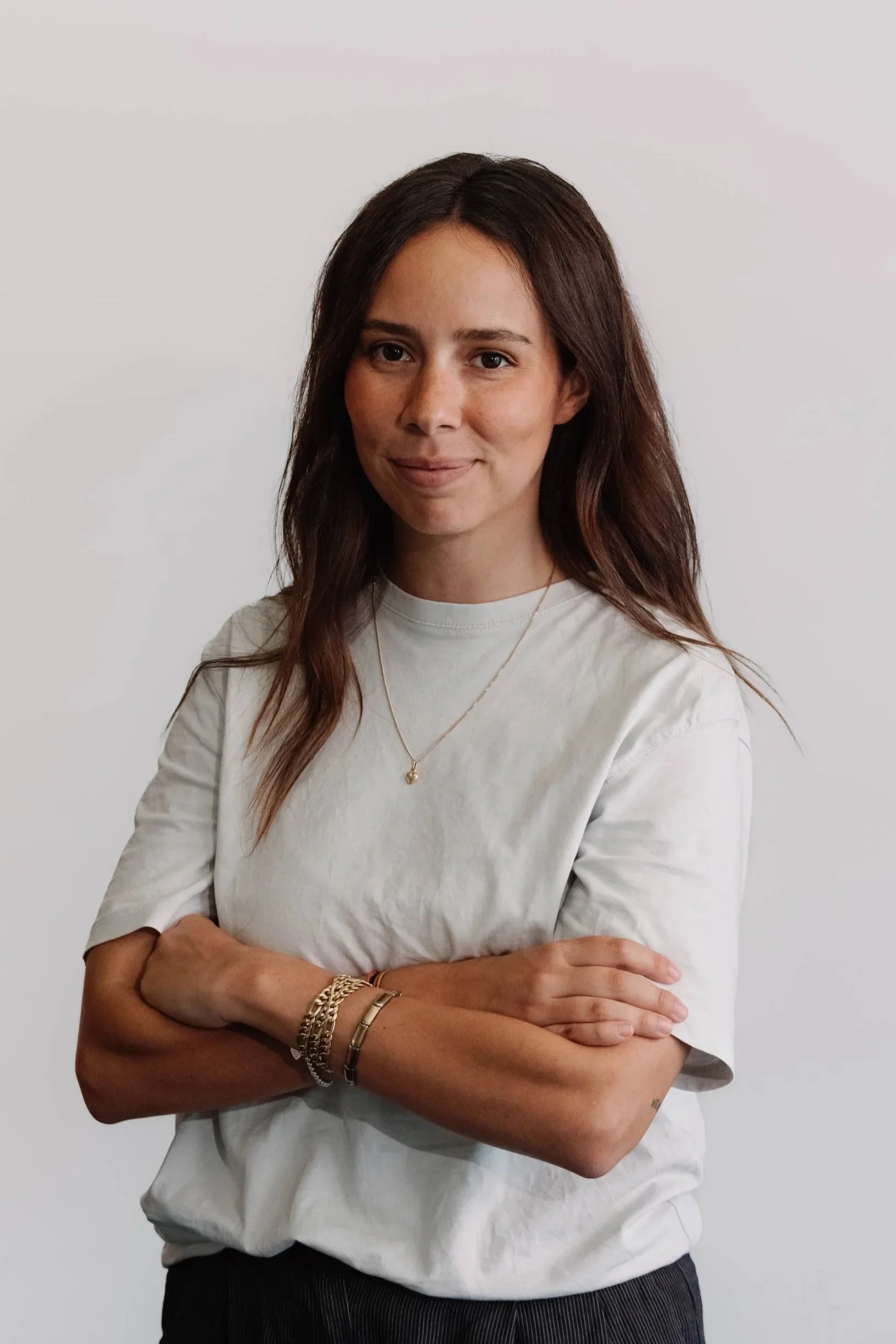 Una donna con capelli marroni, abbigliamento casual, che sorride con le braccia incrociate davanti a uno sfondo bianco.