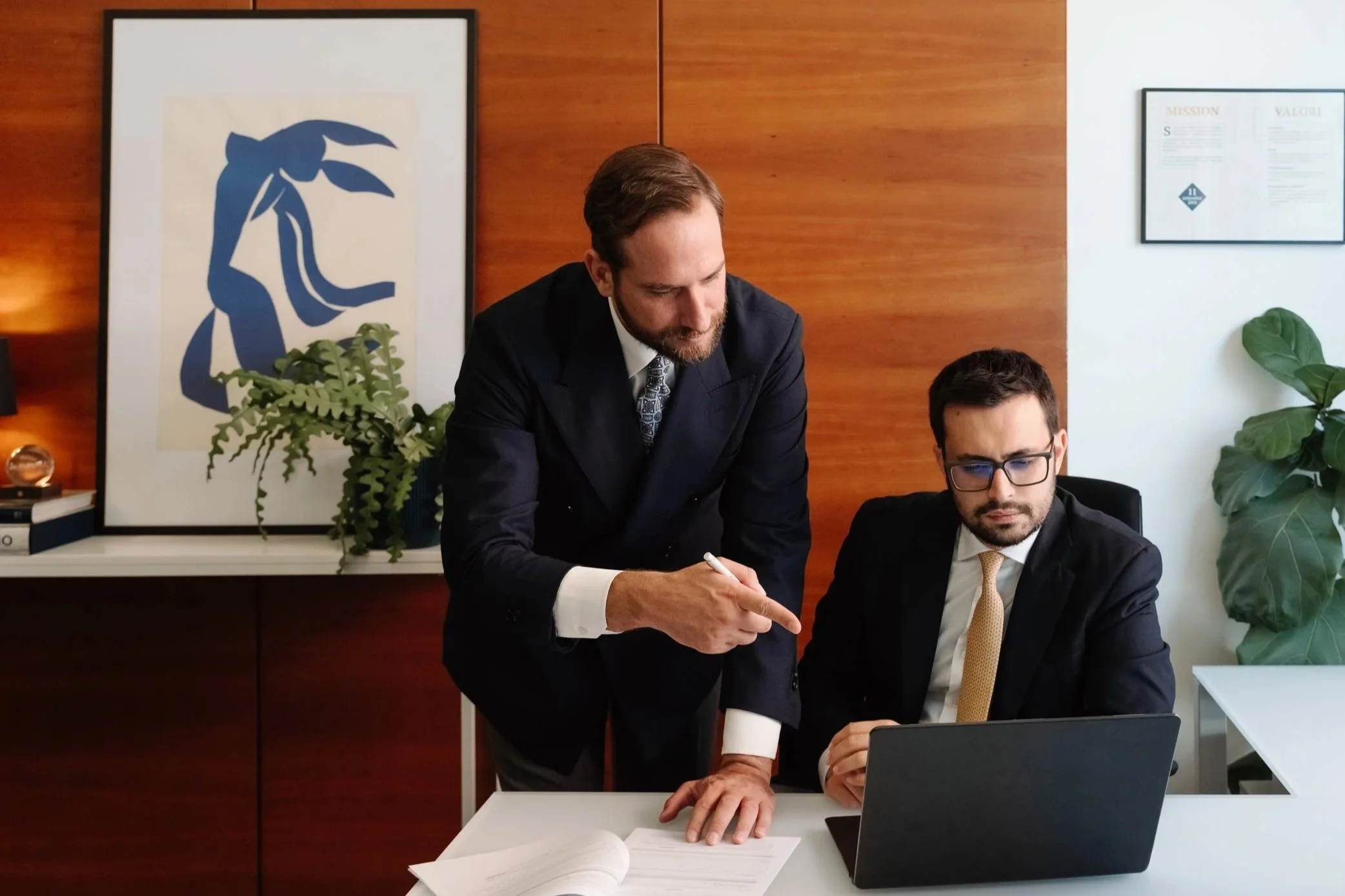 Due uomini in abiti da lavoro discutono davanti a un computer in un ufficio moderno con pareti di legno, piante e quadri art deco.