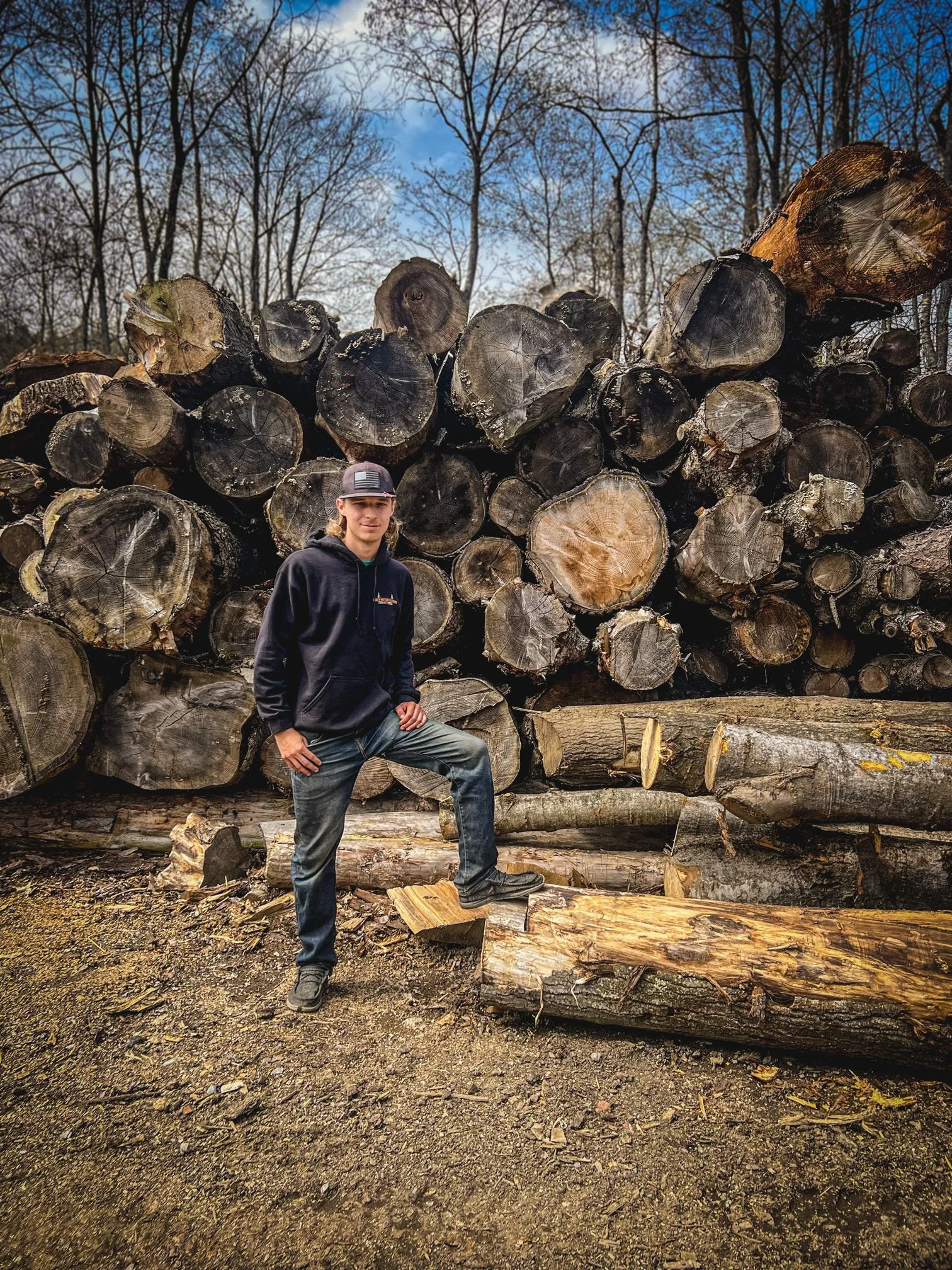 A person stands with one foot on a large fallen log in front of a stack of cut logs in an outdoor wooded area with leafless trees and a partly cloudy sky.