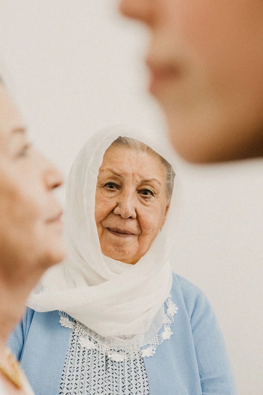 Three people standing together, with an older woman wearing a light headscarf in focus and two others softly out of focus.