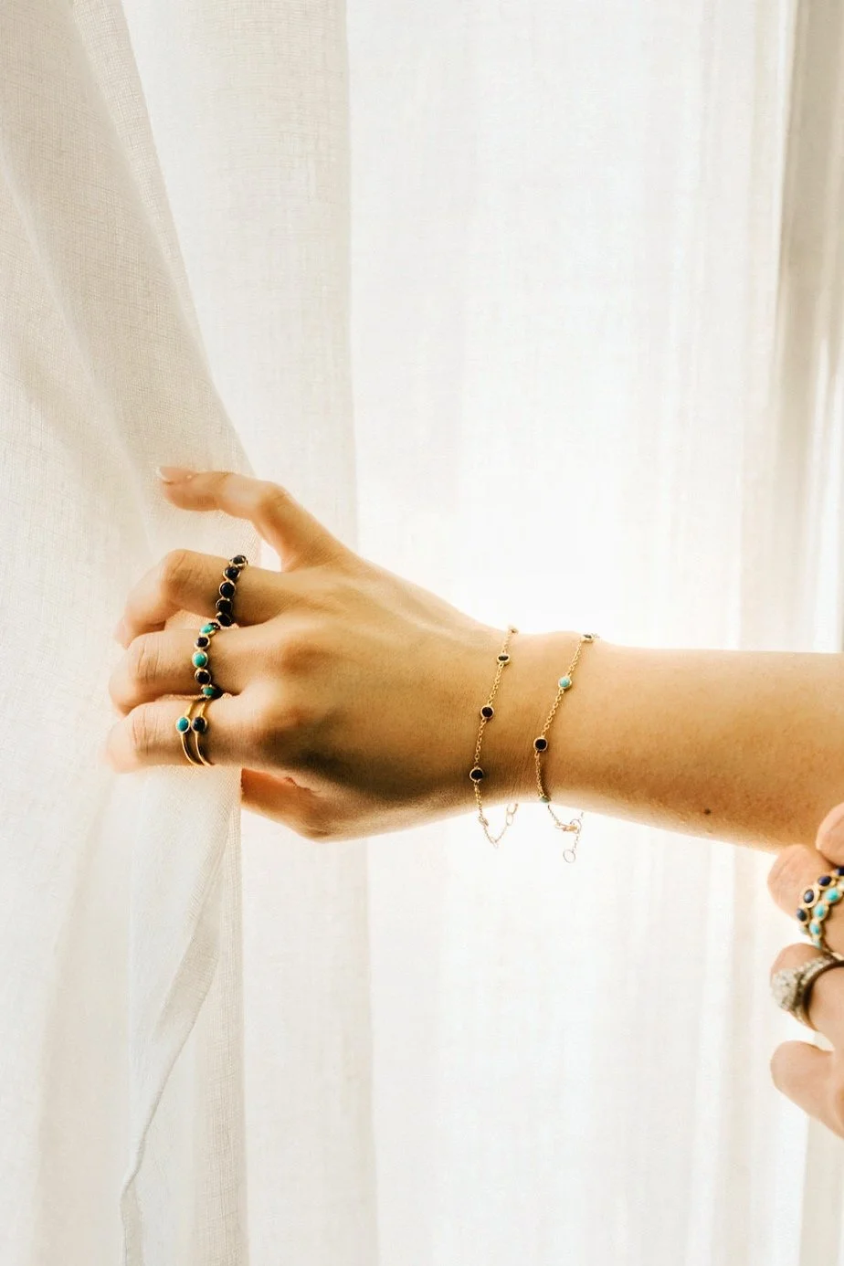 Hand wearing layered gold rings and bracelets gently touching a sheer curtain in soft natural light.