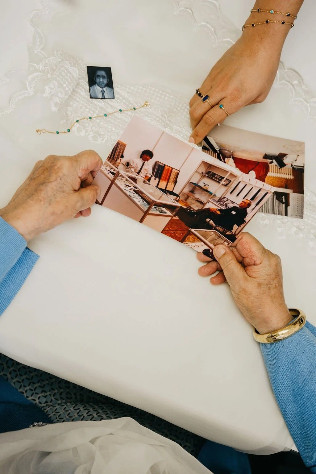 Hands adorned with gold rings and bracelets holding a vintage family photograph on a white lace-covered table, with turquoise jewelry and archival images nearby.
