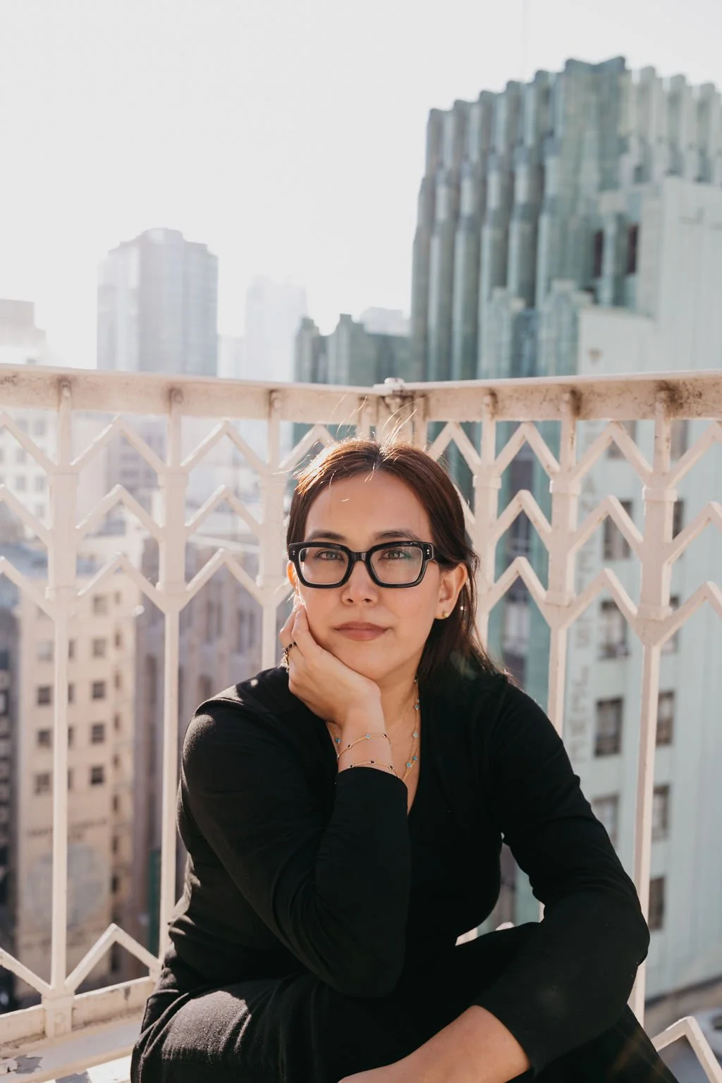 Founder of King Noor LA wearing Paivand necklace, seated on a balcony with Los Angeles city buildings in the background.