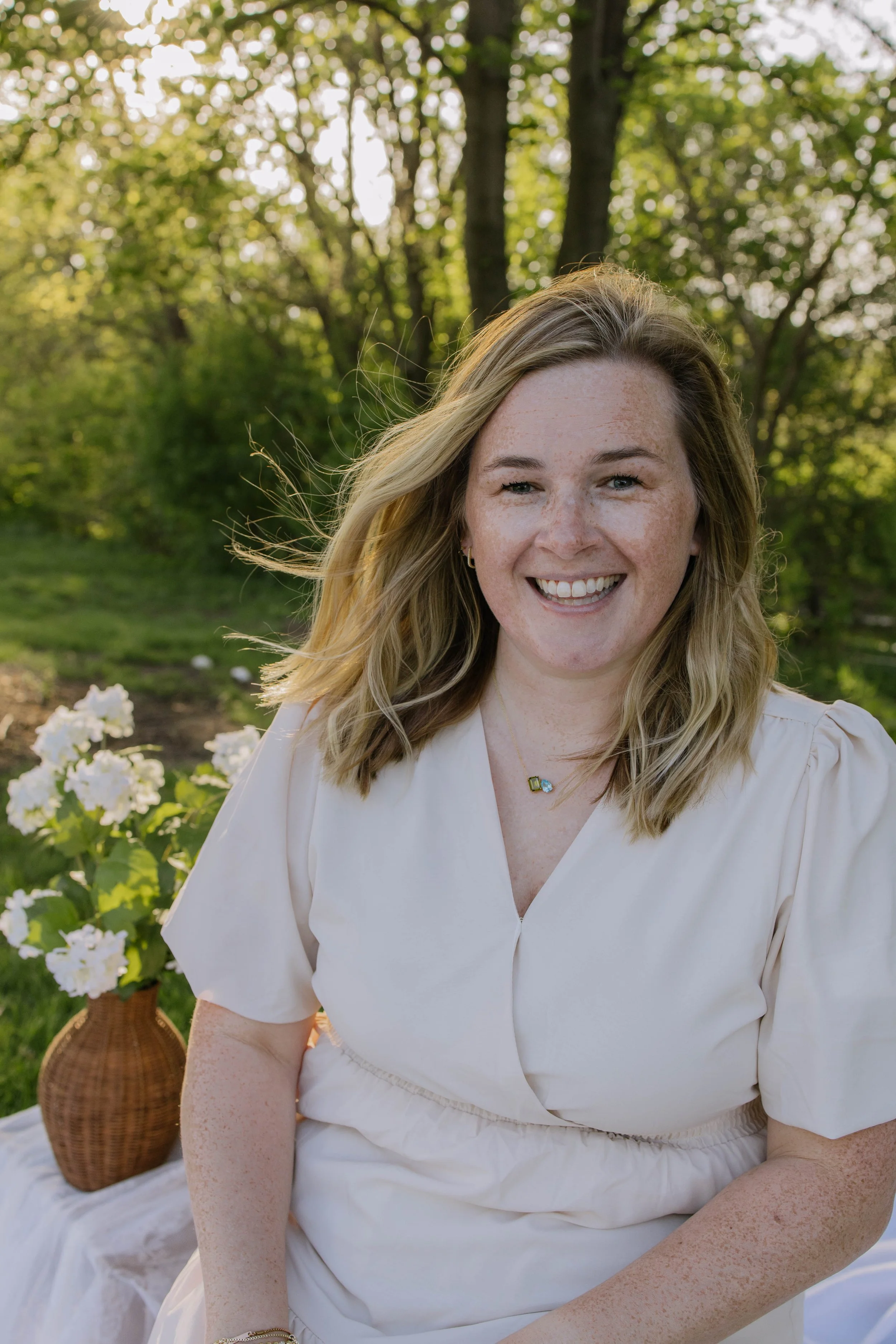 A smiling woman with blonde hair and freckles sitting outdoors, wearing a light-colored dress, surrounded by trees and white flowers.