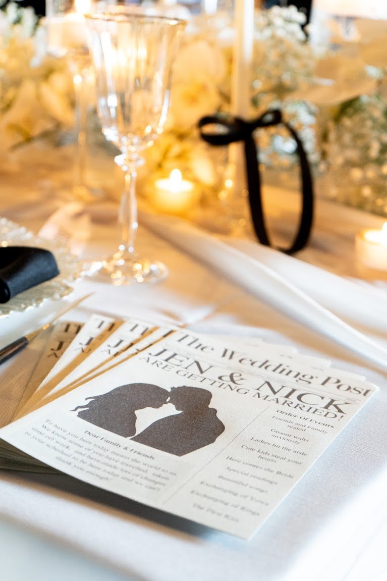 Close-up of a wedding program on a table with glasses, candles, and flowers in the background, featuring a silhouette of a couple and text announcing a marriage.