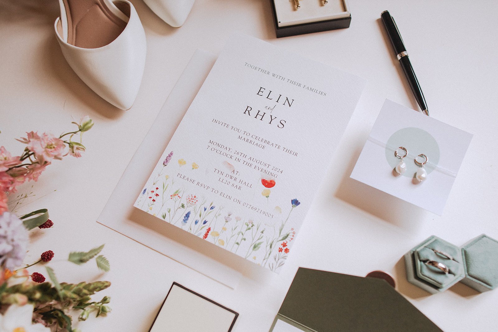 Wedding invitation for Elin and Rhys with floral design, surrounded by accessories like white shoes, jewelry, pen, and flowers.