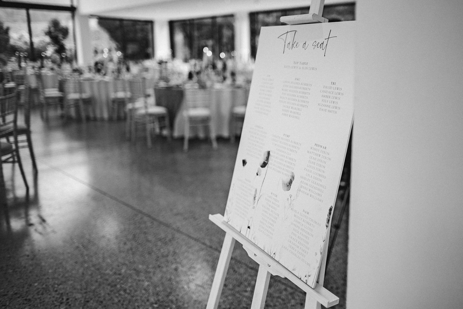Black and white photo of a seating chart on an easel in a reception hall with tables and chairs in the background.