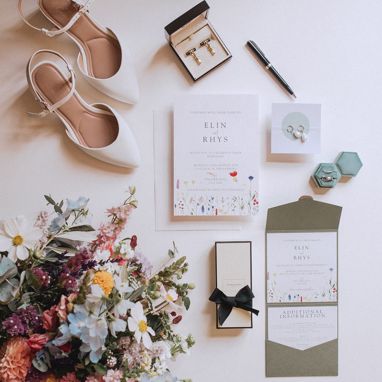 Flat lay of wedding items including white shoes, wedding invitations, a bouquet of flowers, rings, a pen, cufflinks, and a boxed gift.