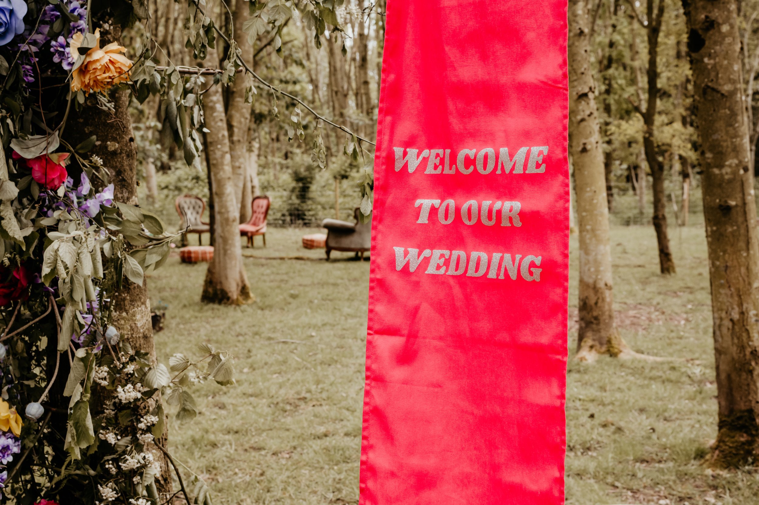 Red wedding banner with 'Welcome to our Wedding' text in outdoor forest setting, with floral decorations and chairs visible in the background.