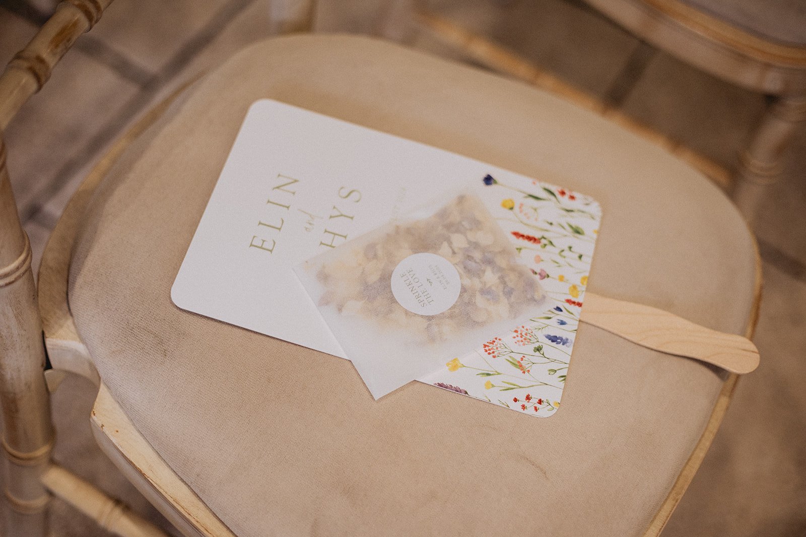 A wedding program and packet of dried petals on a wooden fan with floral design, placed on a beige chair.