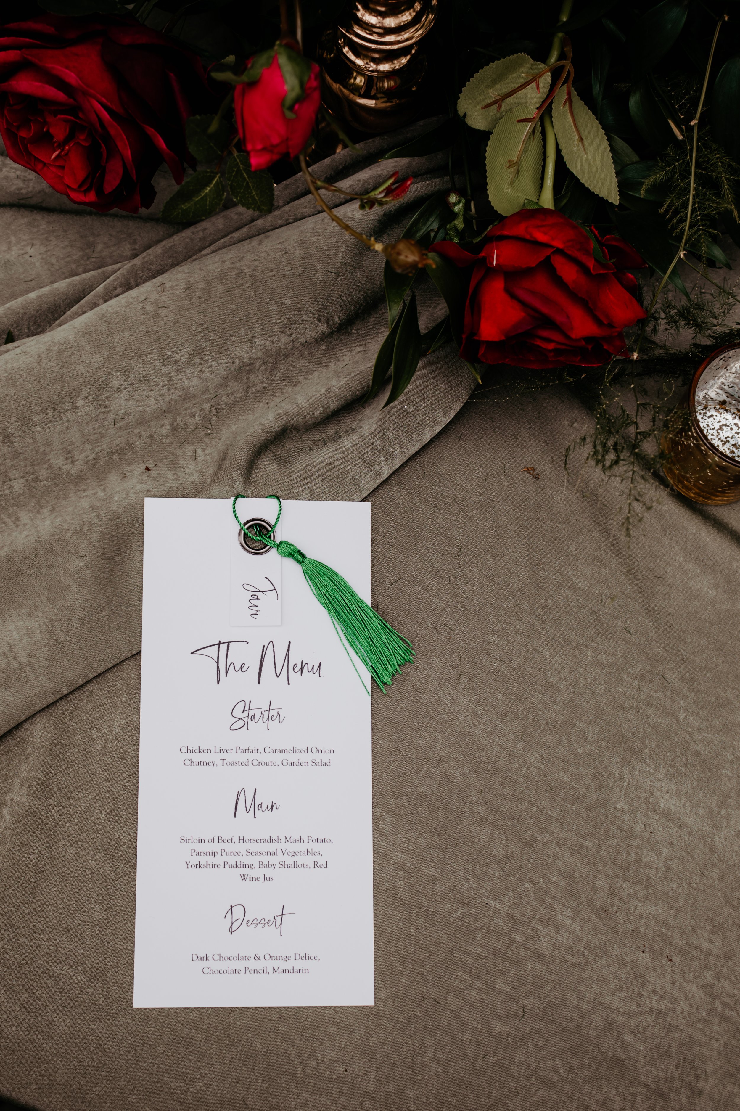 Wedding menu card with green tassel on a gray velvet tablecloth, surrounded by red roses and greenery.