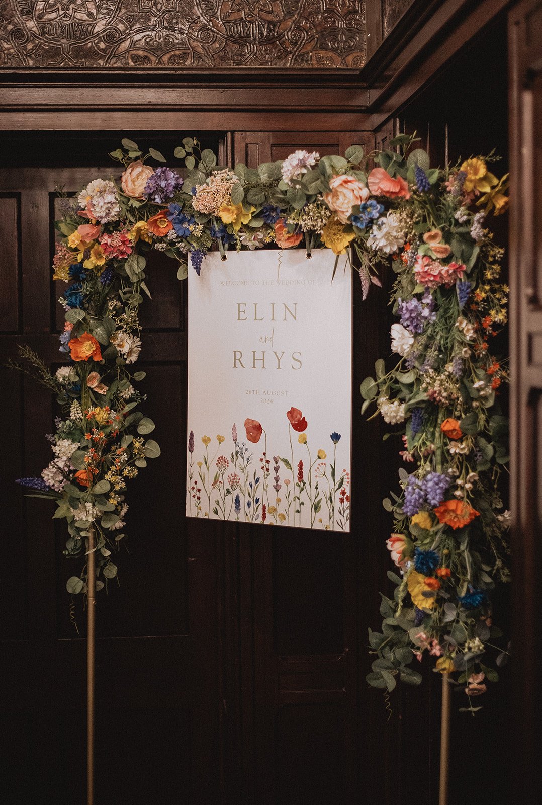 Wedding sign with floral arch; text reads "Welcome to the Wedding of Elin and Rhys, 26th August 2024."