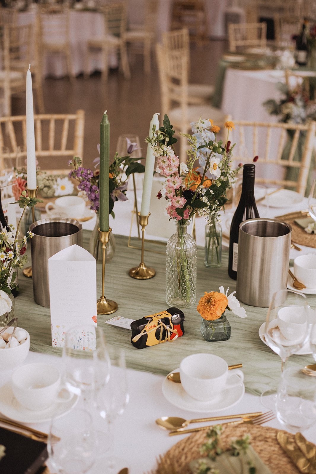 Elegant wedding table setting with floral centerpieces, gold cutlery, green candles, disposable camera, and wine bottle.