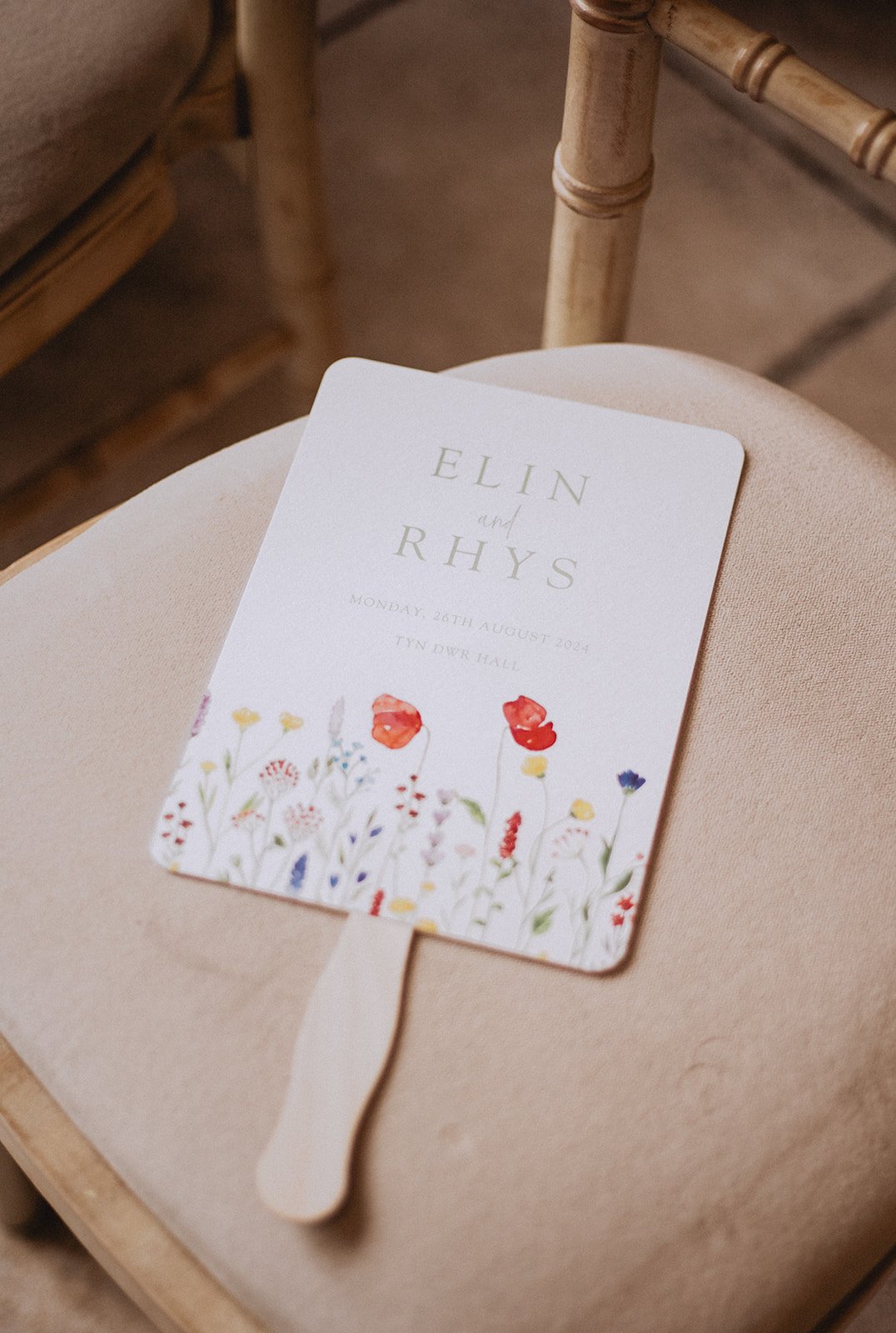 A wedding program on a chair, featuring floral illustrations and the names "Elin and Rhys" with the date and location details.
