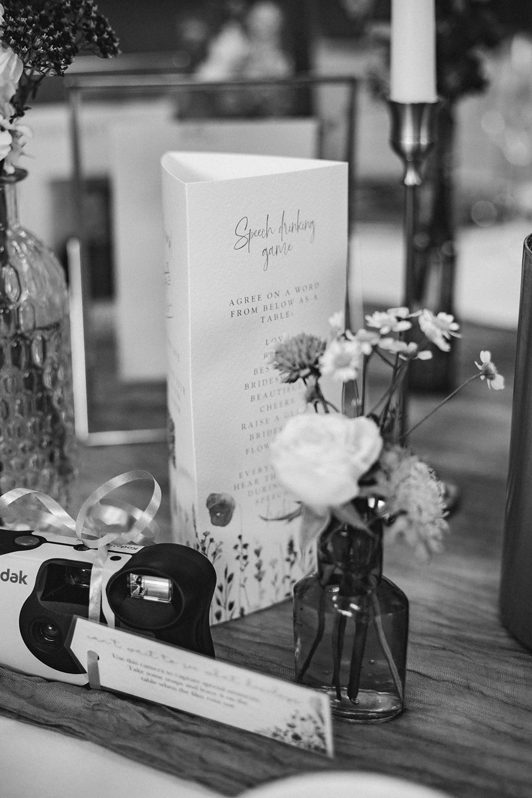 Black and white image of a table setup with a speech drinking game card, a disposable camera with a ribbon, and a small vase of flowers.