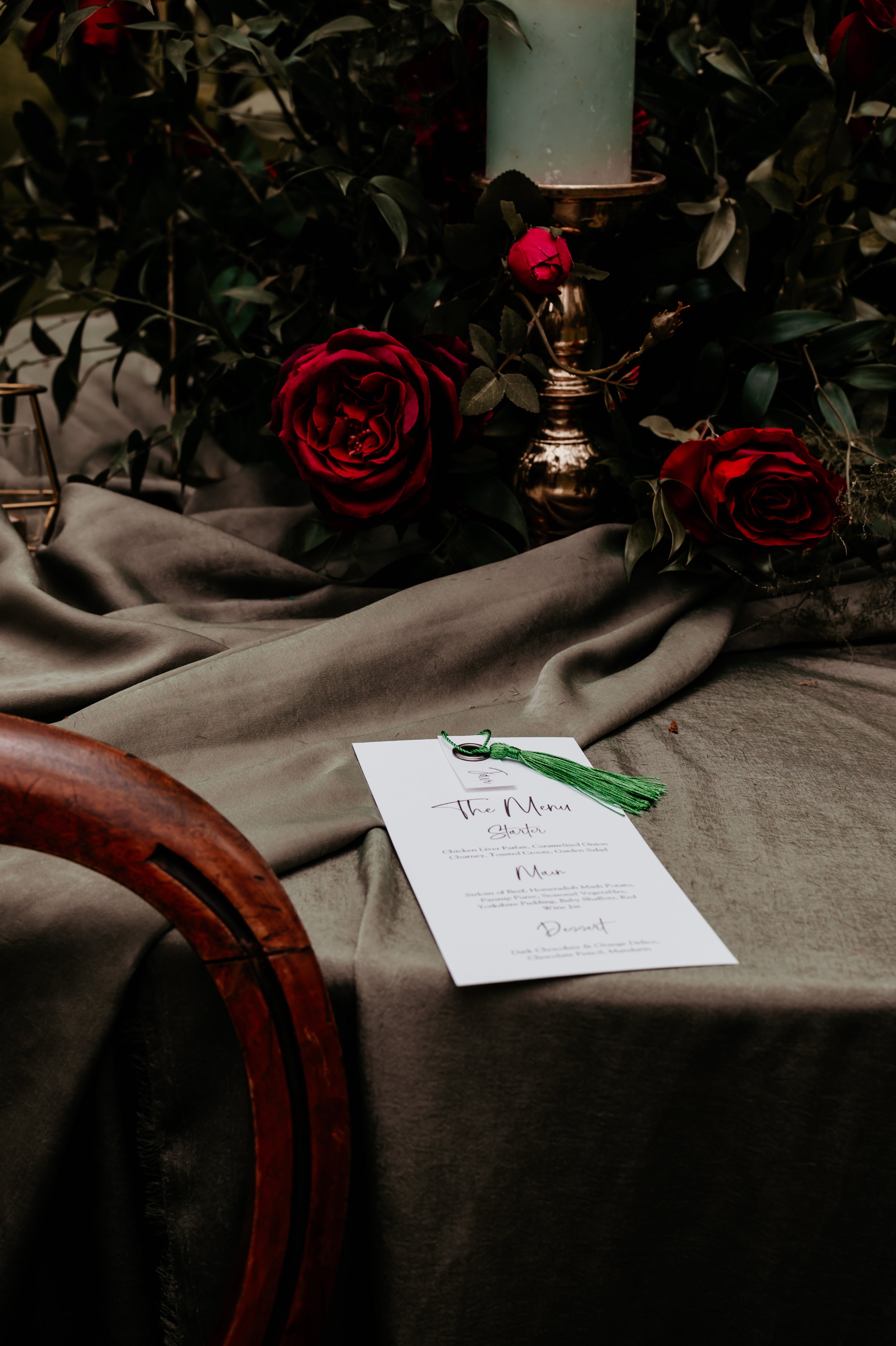 Elegant table setting with red roses, green candle on a brass holder, and a menu card on a gray tablecloth.