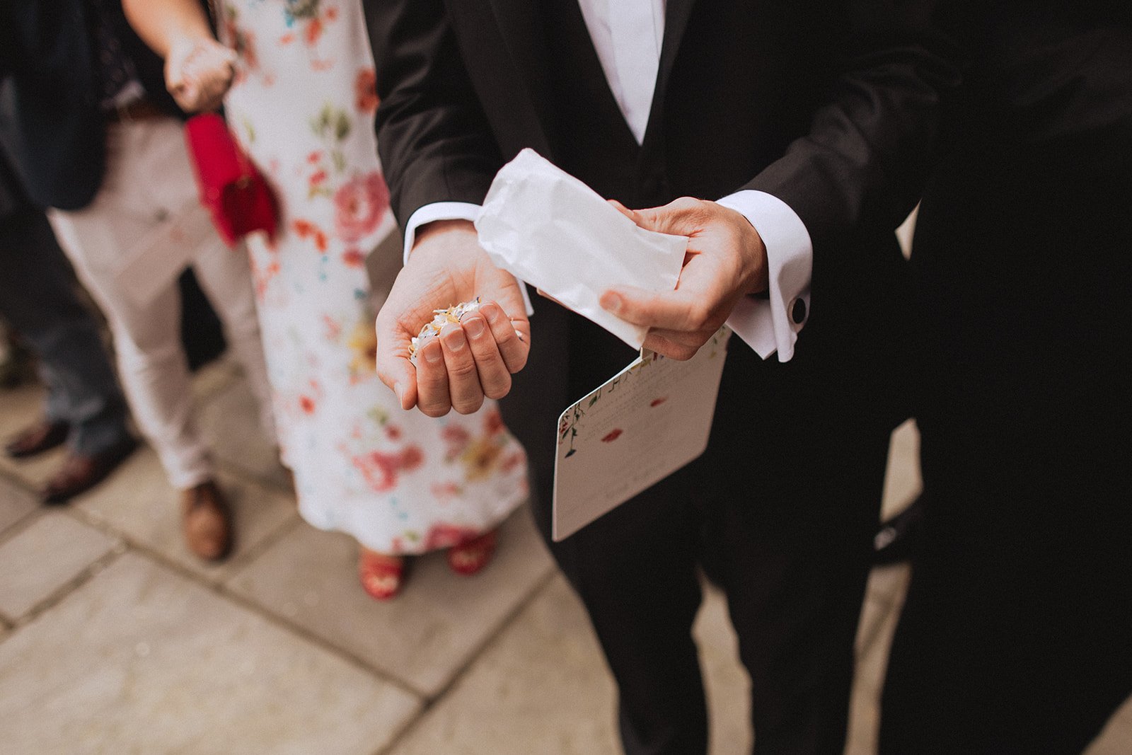 Person holding a small pile of flower petals in one hand, with a white paper bag in the other hand, surrounded by people dressed in formal attire.