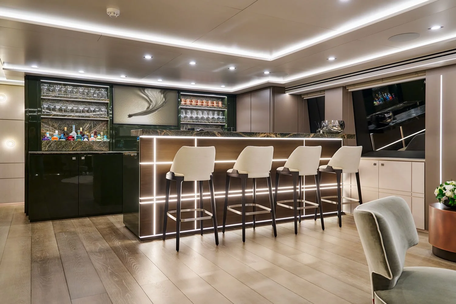 Moderne Bar mit weißen Barhockern, dunkler Barfront mit LED-Beleuchtung, Glasregale mit Spirituosen, Flachbildschirm und dunklen Fensterpaneelen.