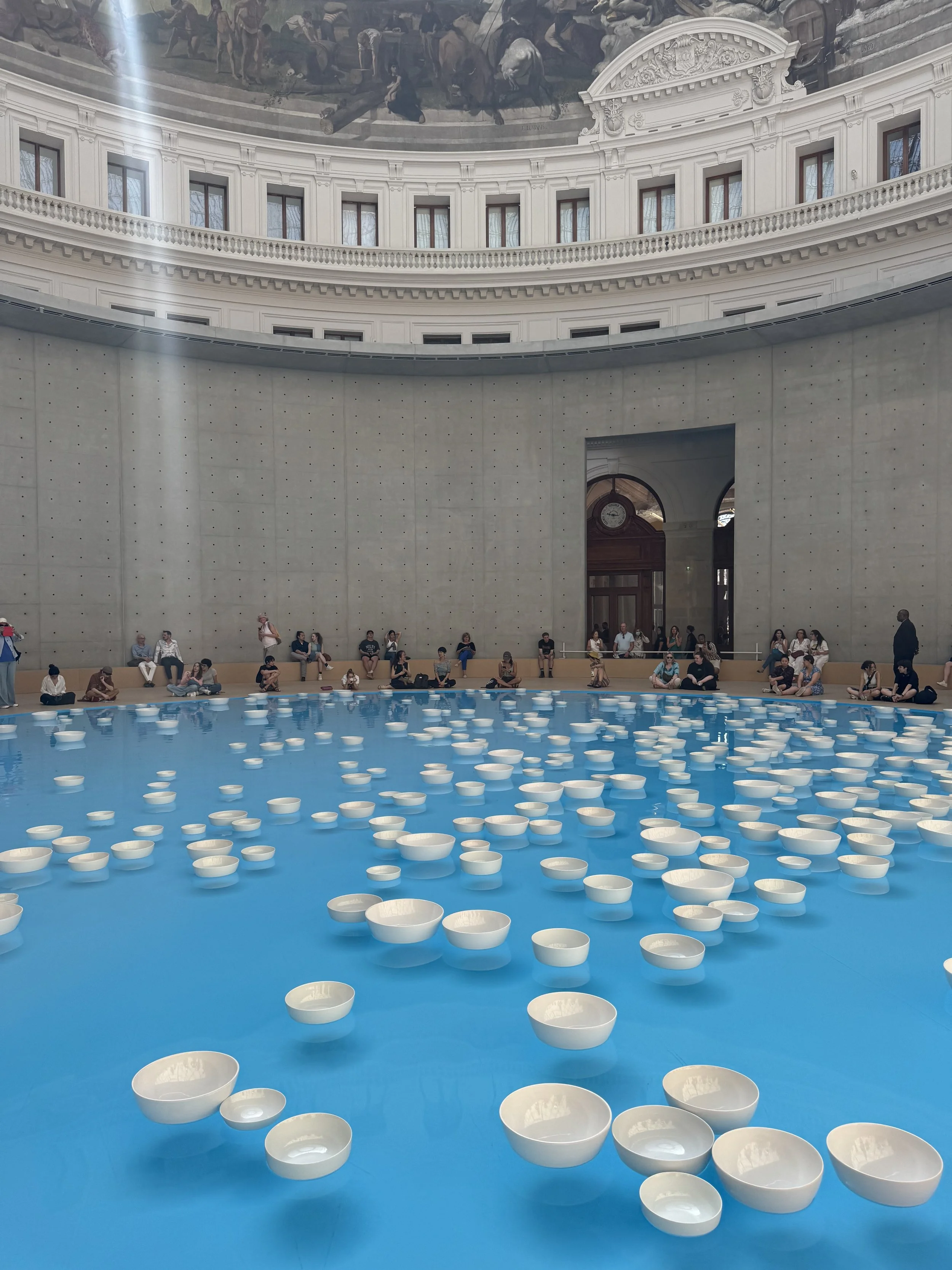 People sitting around a fountain of floating white bowls inside a large, classical building with ornate architectural details and a mural ceiling.