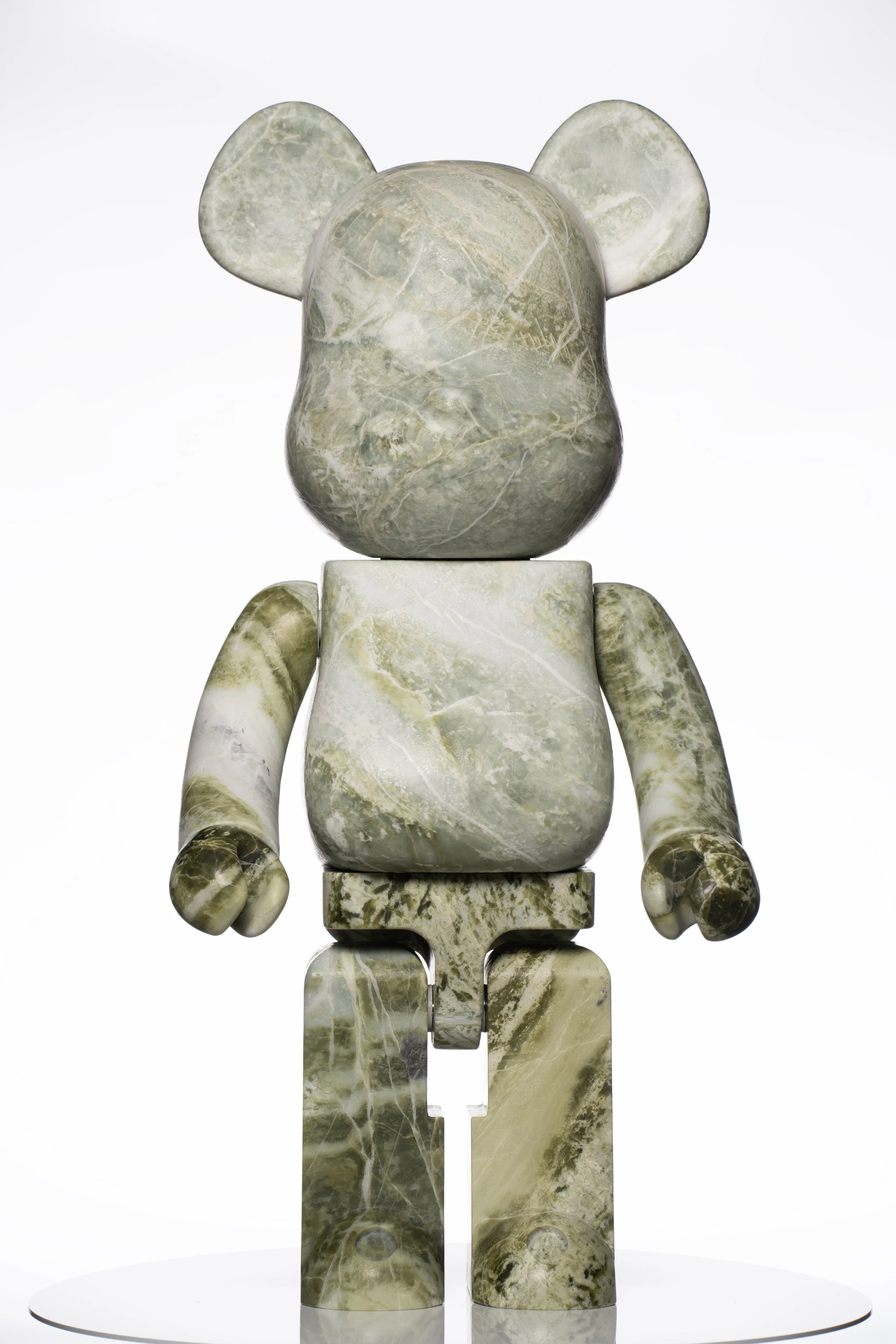 Ein skulpturale Figur eines Teddybären, gefertigt aus grünlich-grauem Marmor, bestehend aus mehreren segmentierten Teilen, auf einer weißen runden Basis.